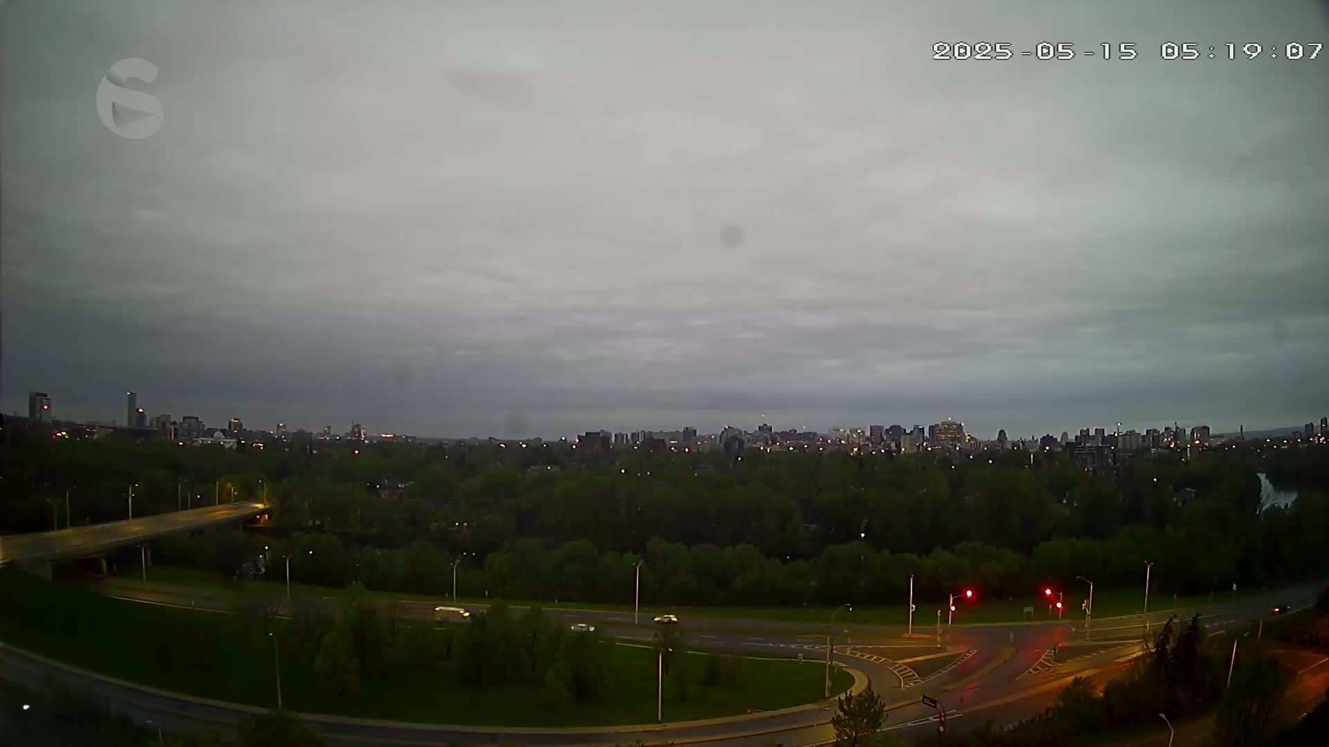 An overcast sky hangs over a city skyline visible beyond a large, tree-filled park and a highway interchange at dusk.
