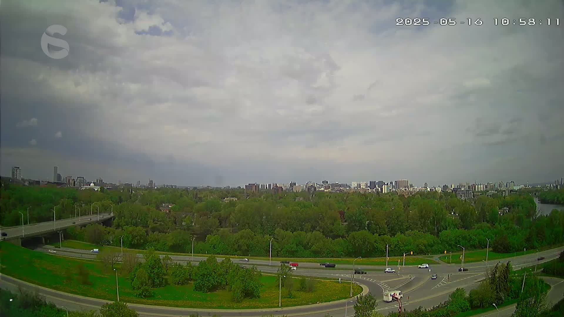An overcast sky overlooks a highway interchange surrounded by lush green trees and a distant cityscape.