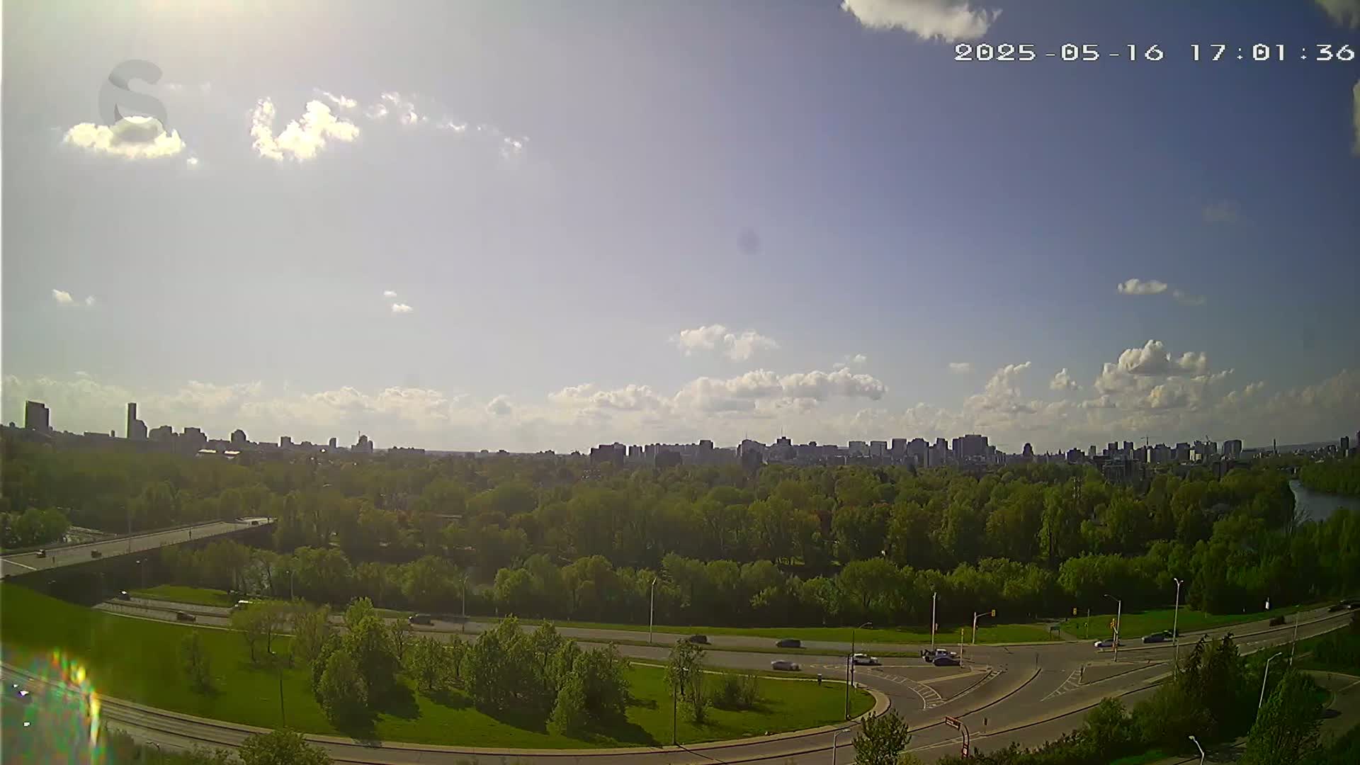 A sunny day with scattered clouds reveals a cityscape beyond a green park and curved roadway.