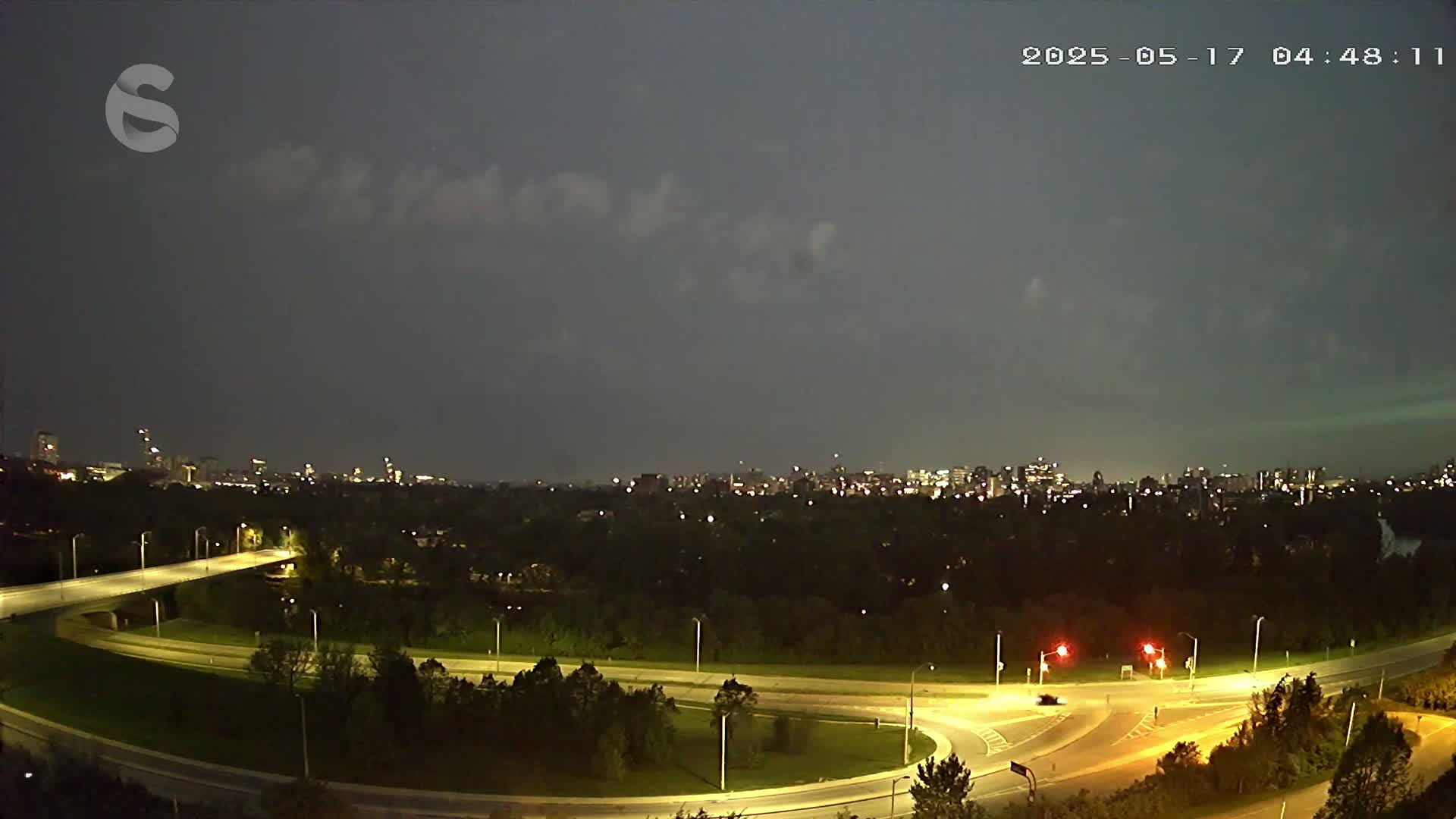 A nighttime view of a city skyline across a highway interchange under a mostly cloudy sky.