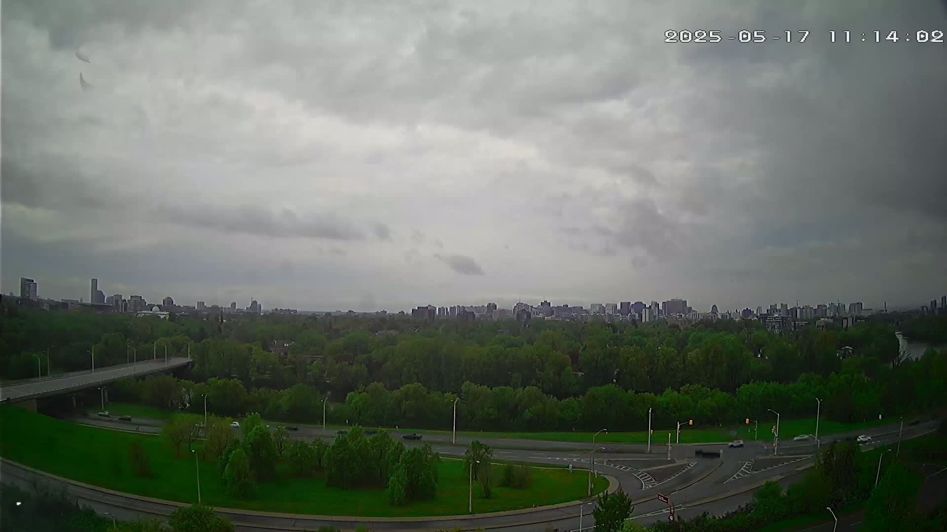 A city skyline is visible in the distance beyond a lush green park with roads and a bridge under an overcast sky.