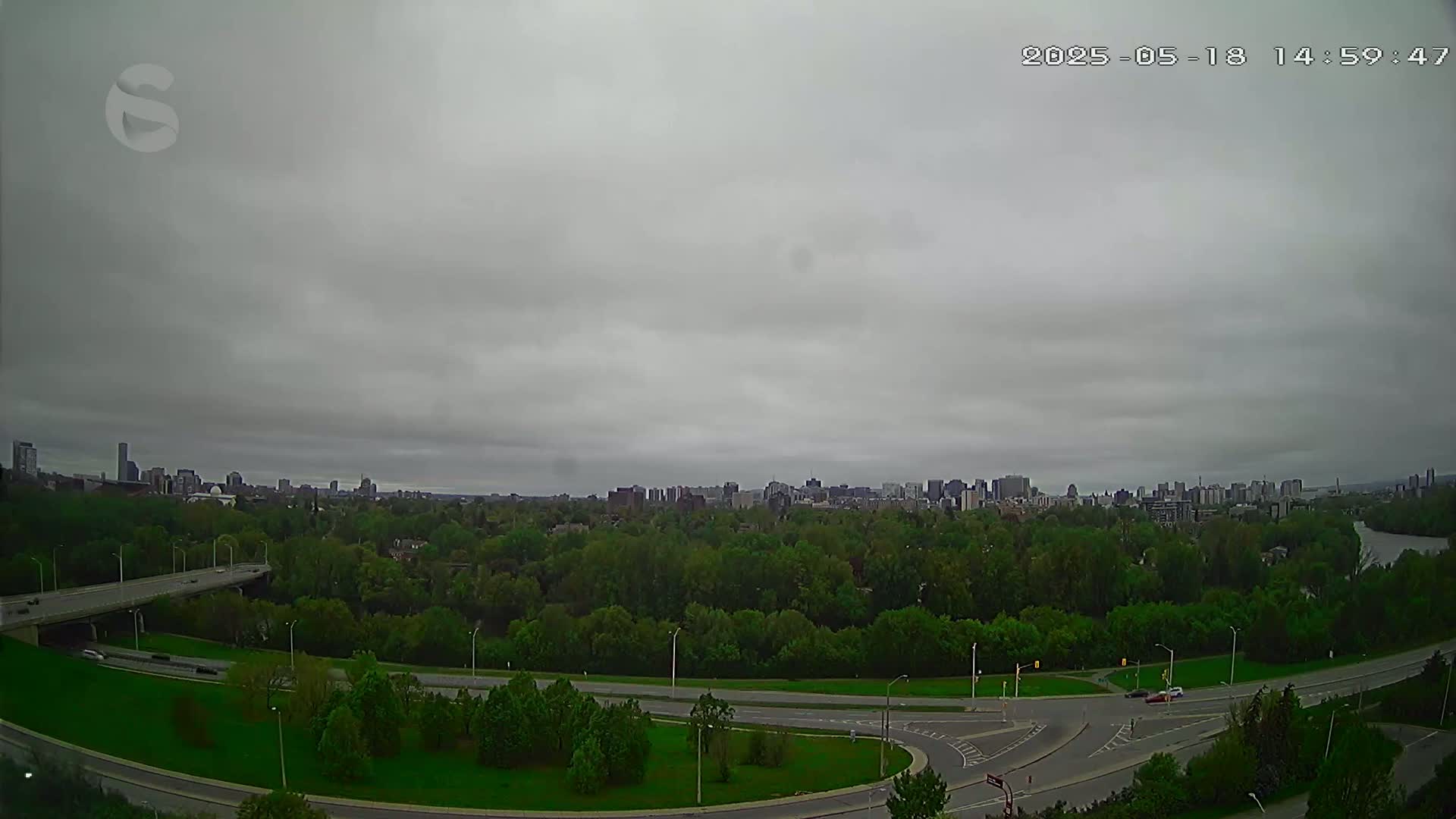A city skyline is visible in the distance under a cloudy sky, with a highway and green space in the foreground.