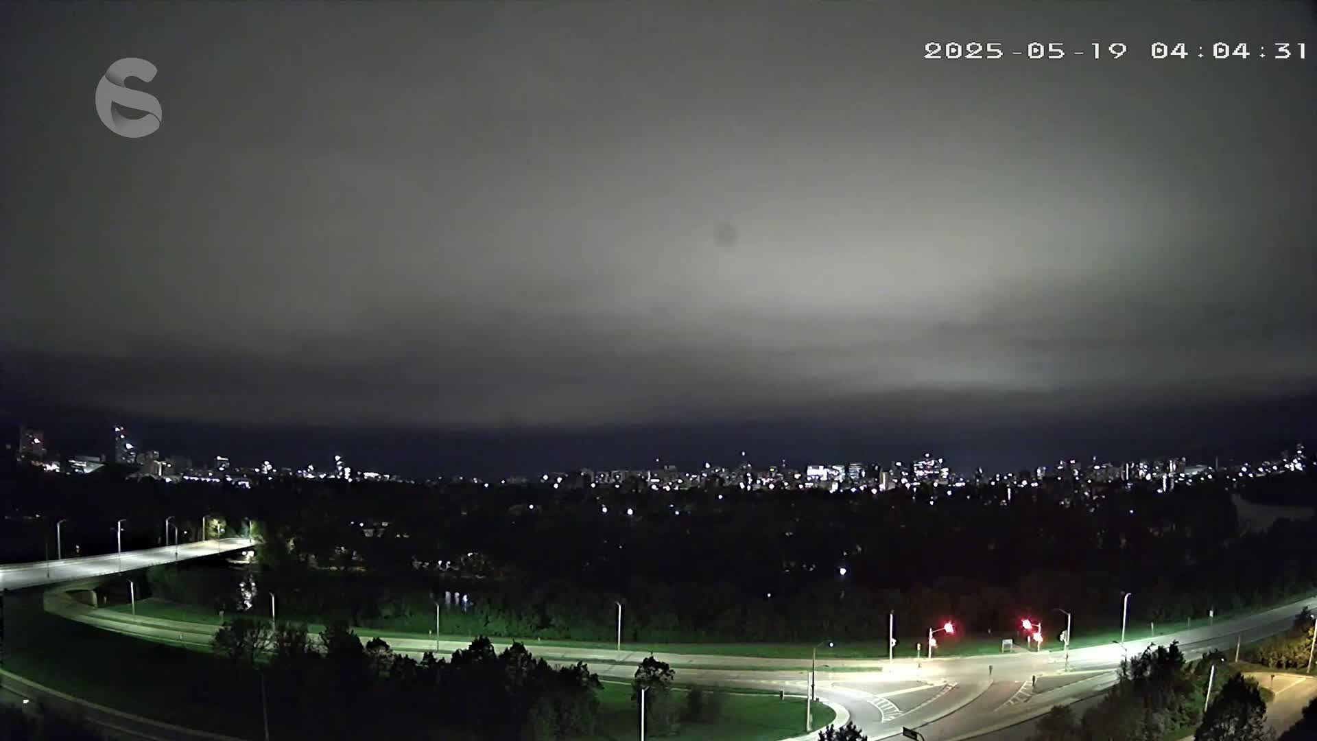 A nighttime view of a city skyline across a wooded area and a highway interchange under an overcast sky.