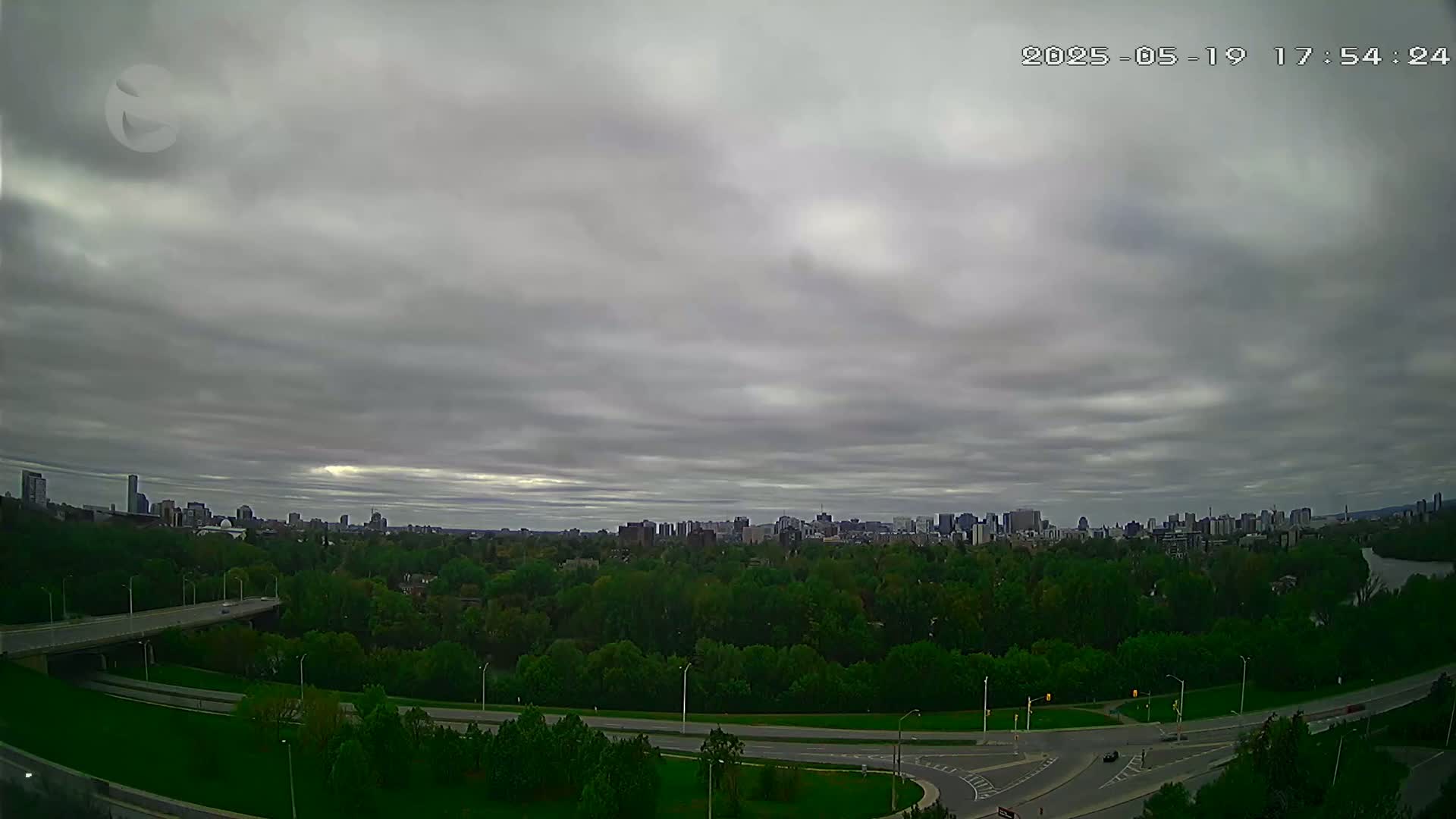 An overcast sky hangs over a city skyline partially obscured by a lush green expanse of trees and winding roads.