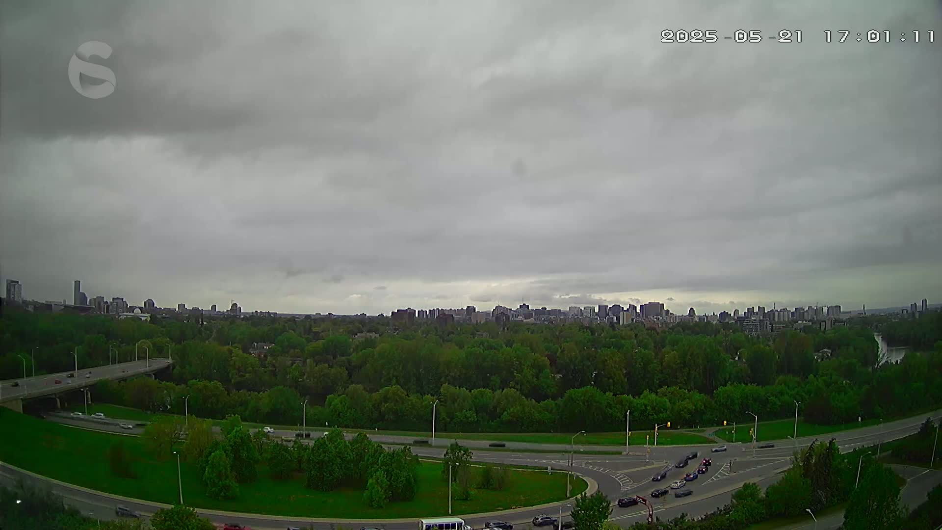 A city skyline is visible in the distance beyond a highway and lush green parkland under an overcast sky.