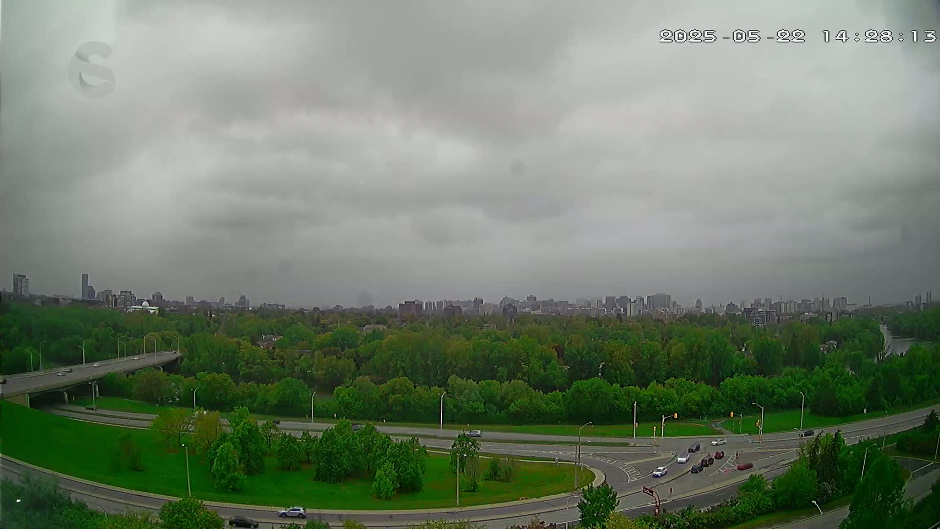 A highway interchange curves through a lush green park, with a distant cityscape visible under an overcast sky.