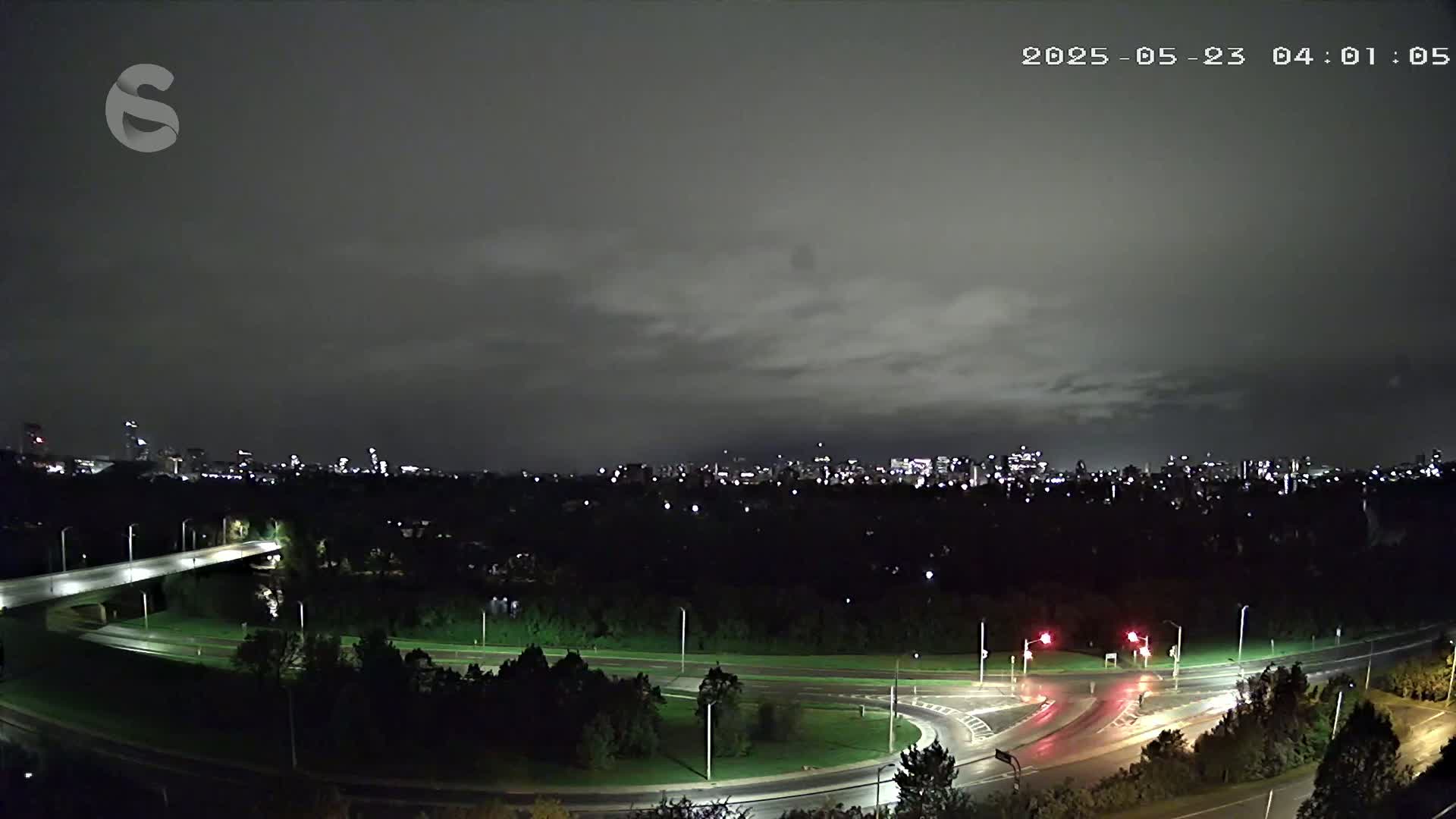 A nighttime, overcast view of a city skyline beyond a highway interchange and a wooded area, with wet roads.