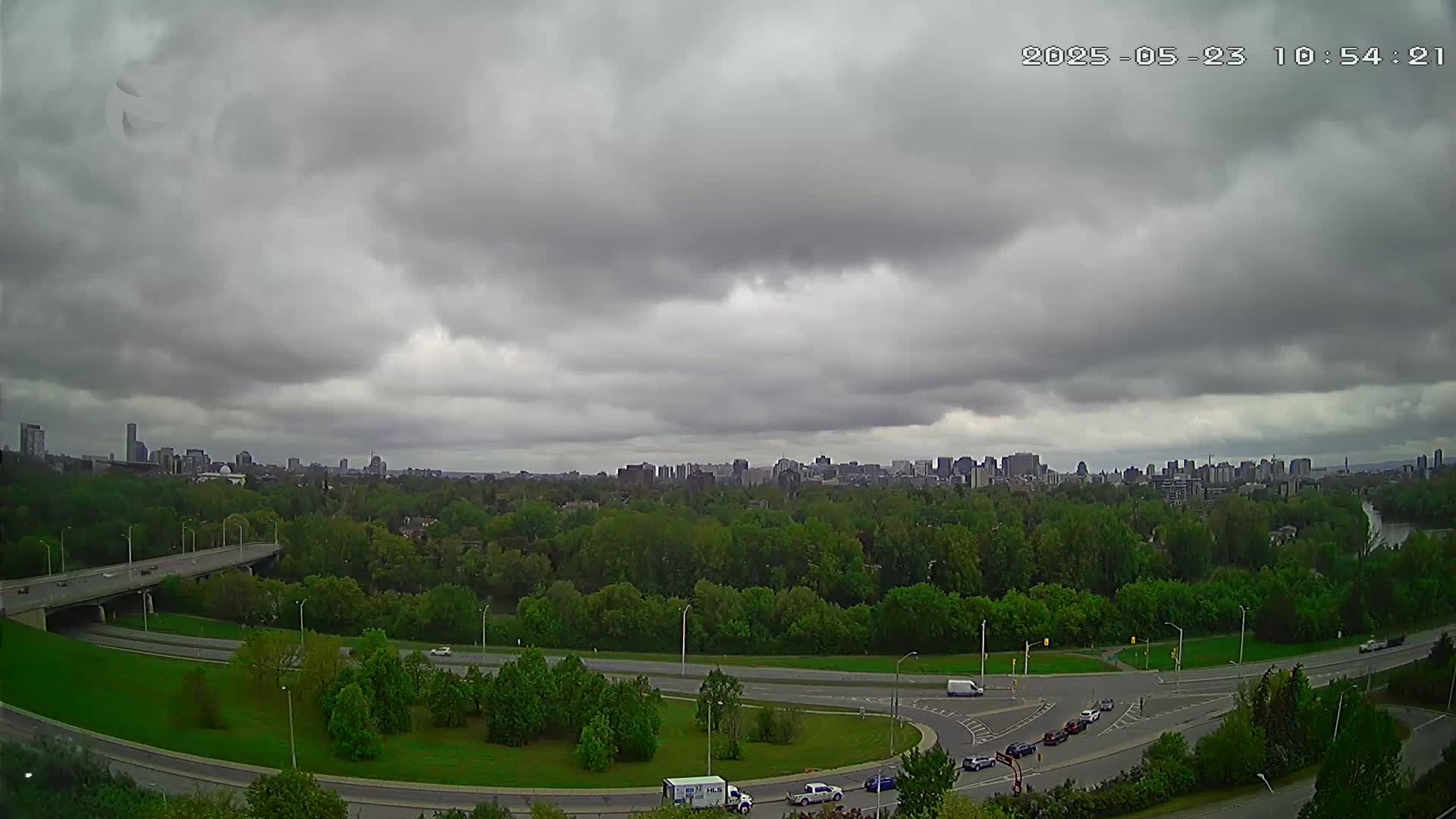 A highway interchange curves through a green, tree-lined area under an overcast sky, with a city skyline visible in the distance.