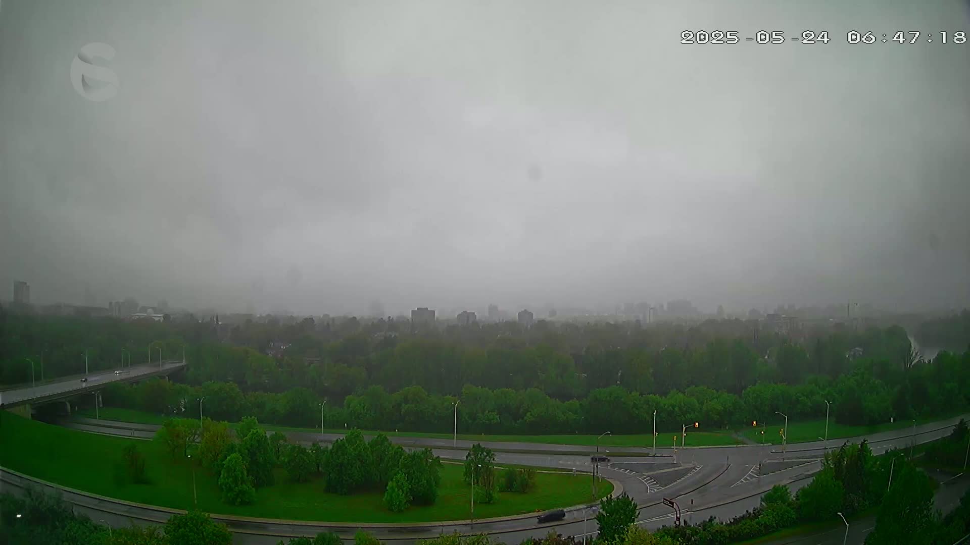 A highway overpass and a large roundabout are visible through a hazy rain over a lush green landscape, with a distant cityscape barely visible in the background.