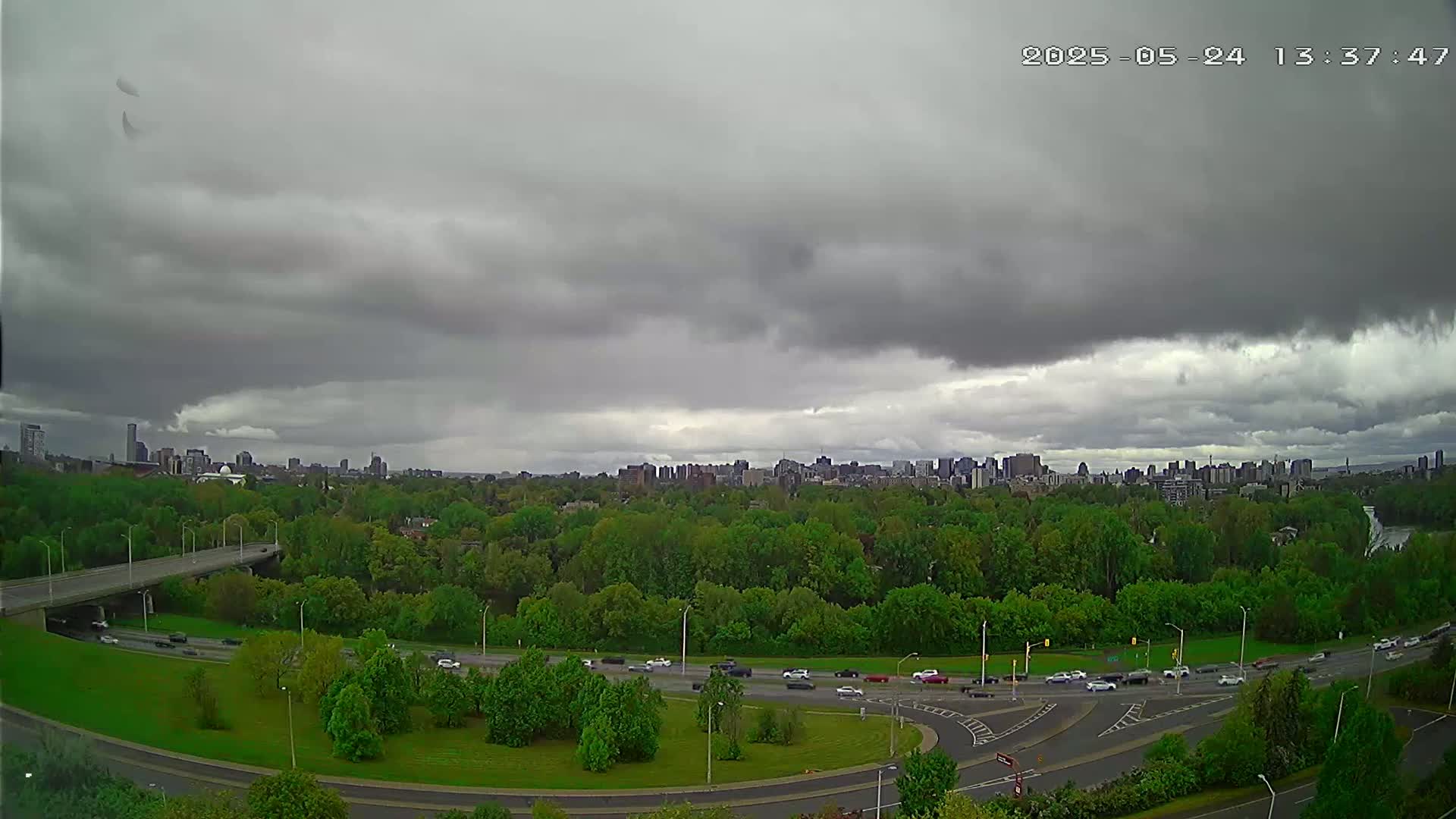 A city skyline is visible in the distance beyond a lush green park and highway with overcast skies.