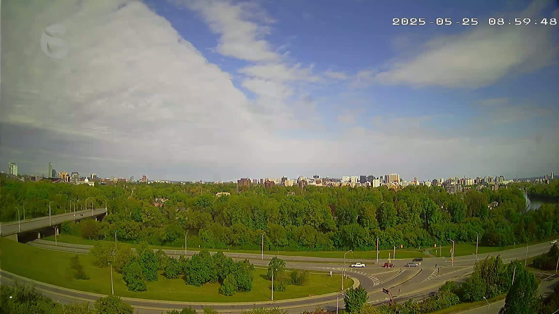 A partly cloudy sky overlooks a highway interchange and a cityscape nestled in a lush green forest.