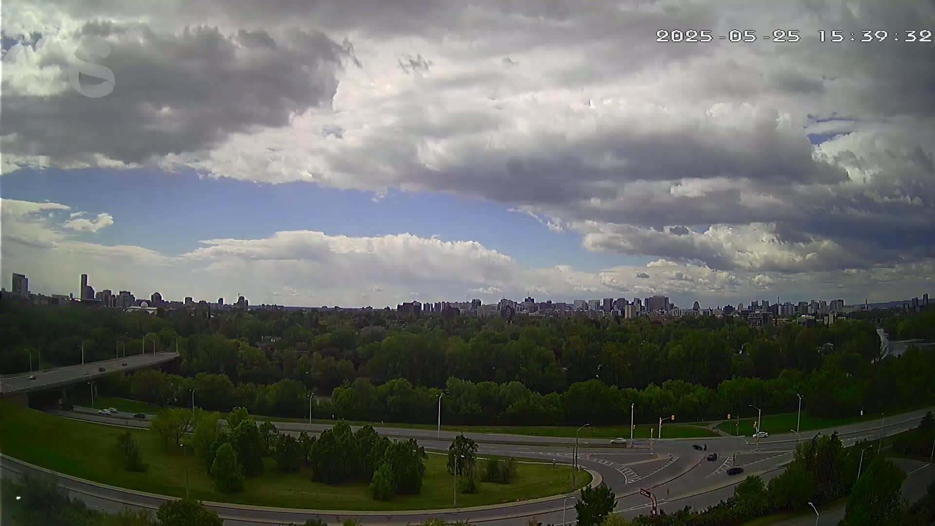A partly cloudy sky overlooks a highway interchange and a city skyline in the distance.