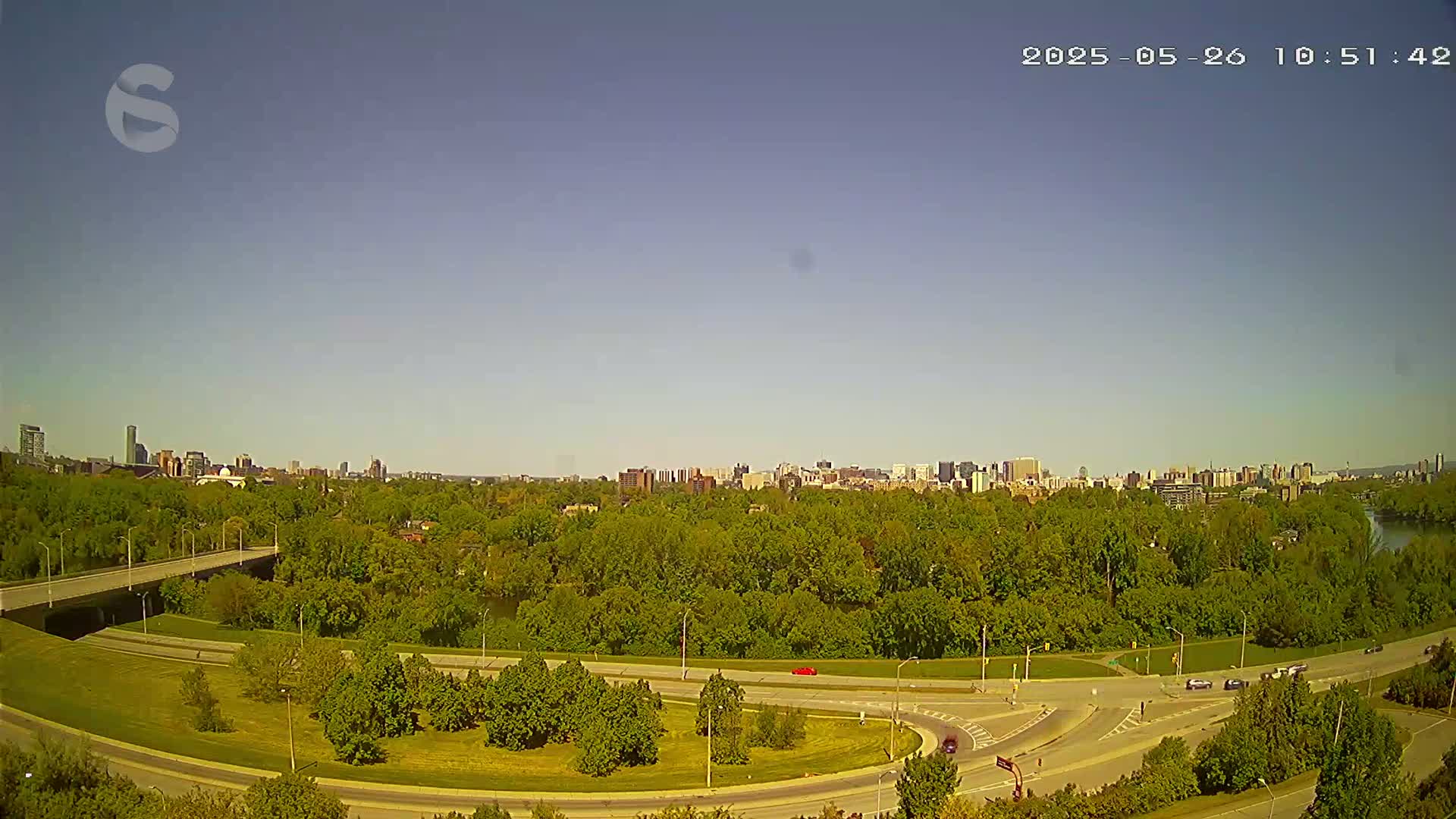 A highway curves through a lush green landscape under a clear blue sky, with a distant city skyline visible in the background.
