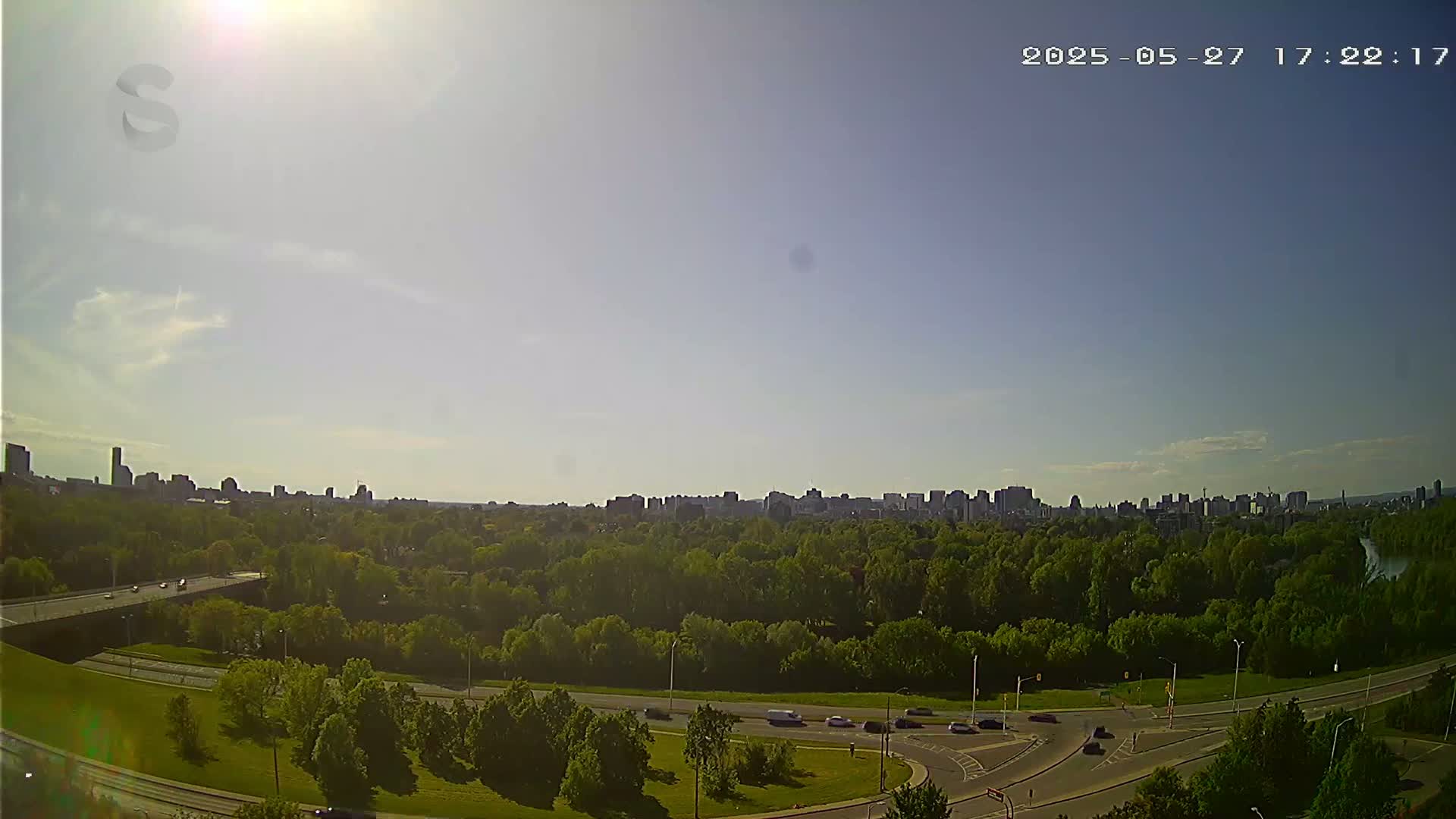 A sunny cityscape is visible in the distance beyond a green expanse of trees, roads, and a highway overpass.