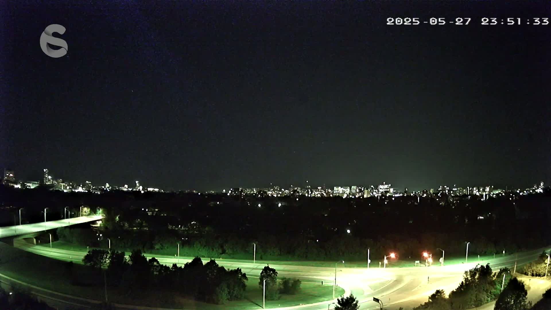 A nighttime view of a city skyline across a highway interchange and wooded area under a clear, dark sky.