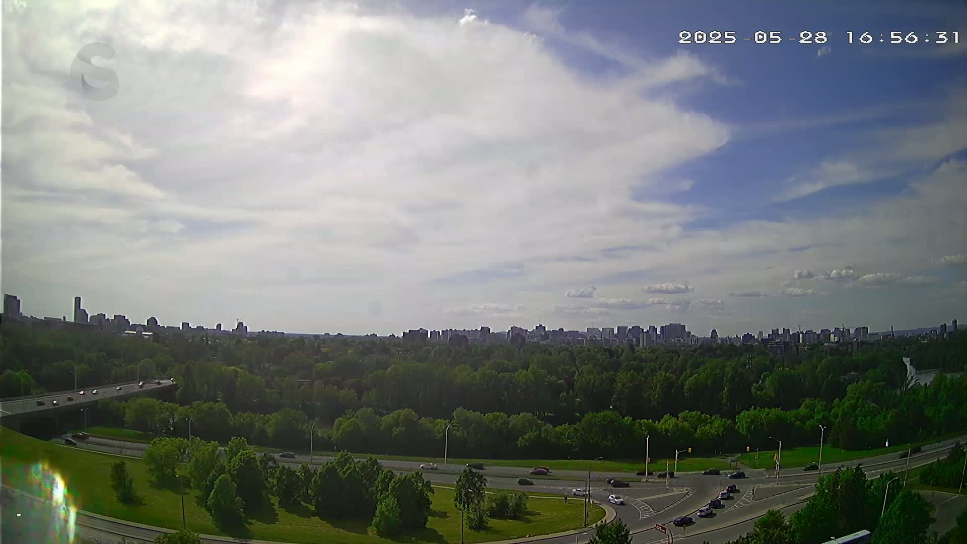 A partly cloudy sky overlooks a city skyline beyond a sprawling green park with several roads and vehicles visible.