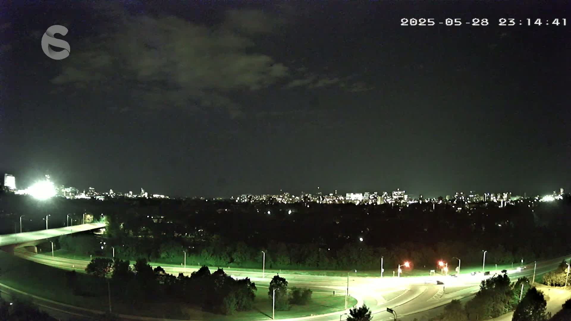 A nighttime view of a city skyline across a tree-lined highway intersection under a partly cloudy sky.