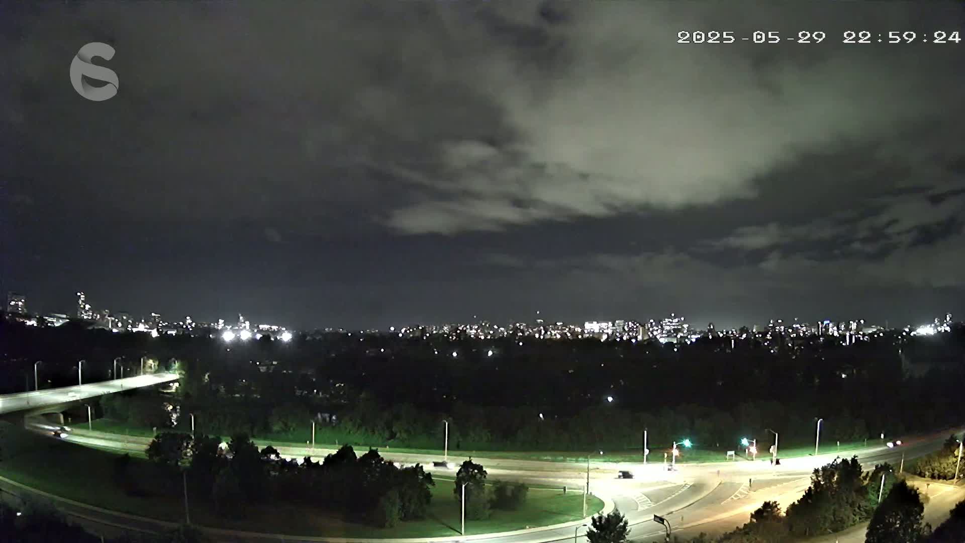 A nighttime view of a city skyline beyond a dark, tree-covered area, with a highway curving through the foreground under a mostly cloudy sky.