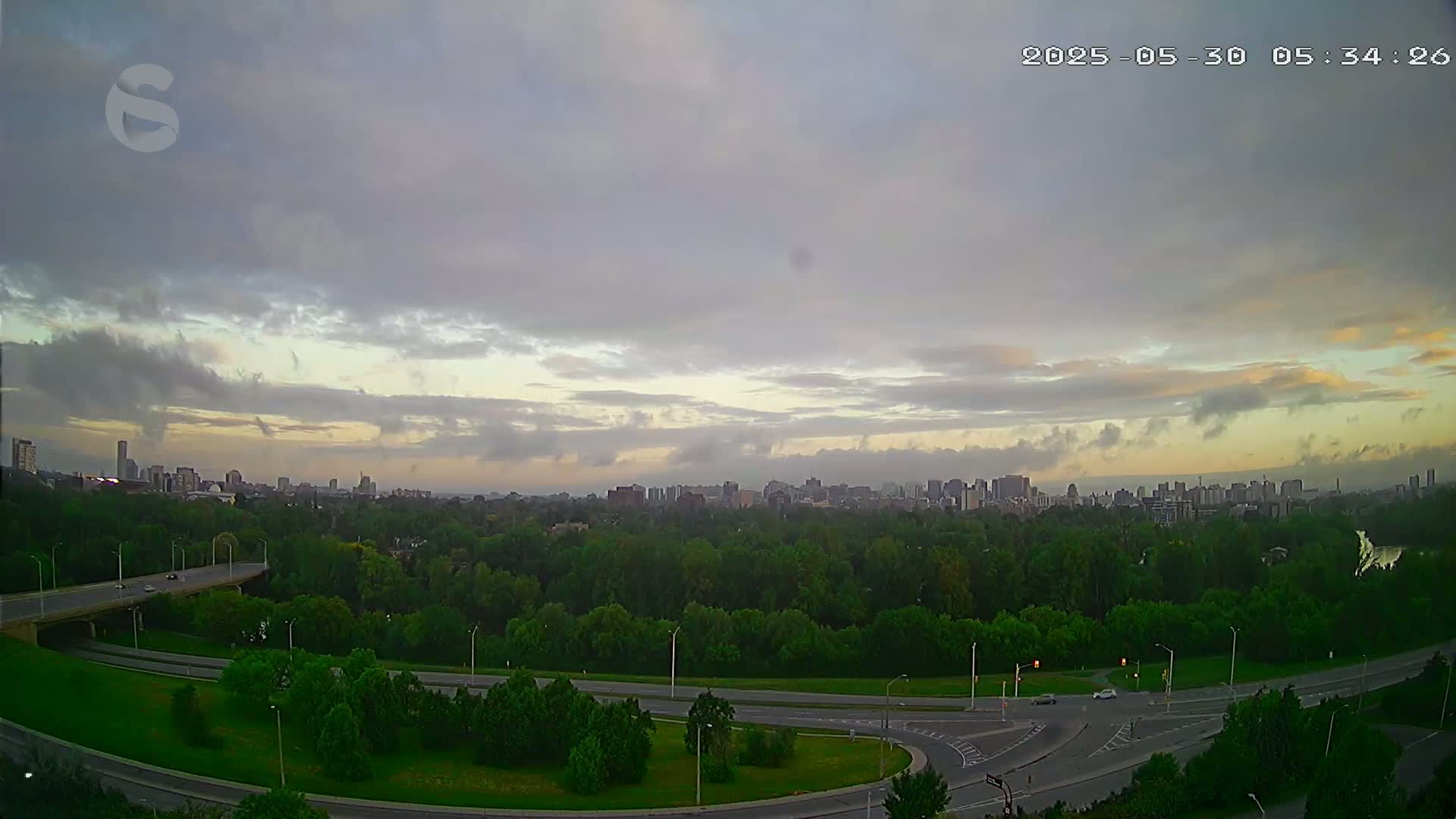 A city skyline is visible in the distance beyond a vast green expanse of trees and a curving highway under a mostly cloudy, sunrise sky.