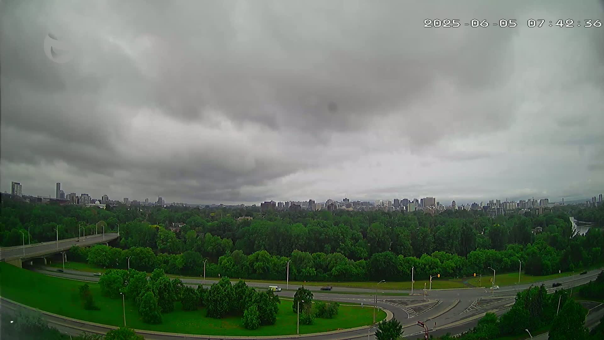 A city skyline is visible in the distance beyond a vast expanse of green trees and a curving highway under an overcast sky.