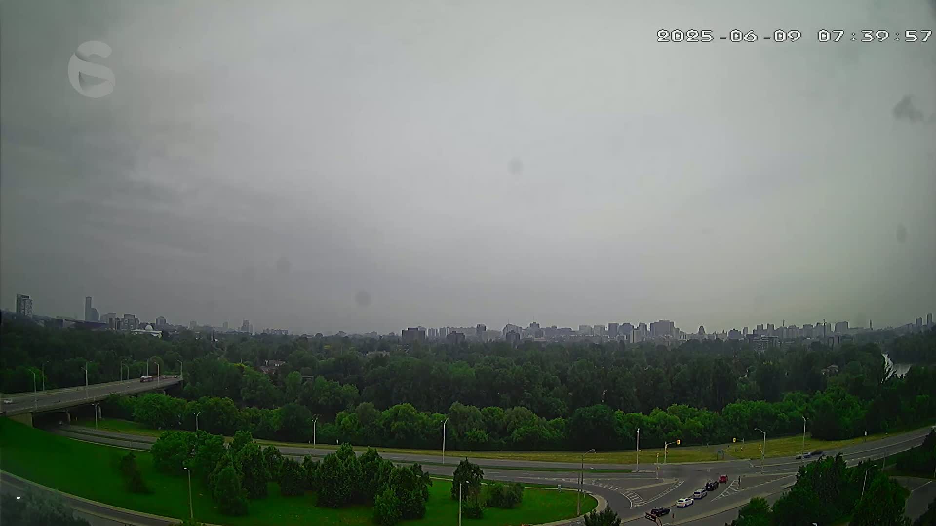 A hazy, overcast sky hangs over a highway system curving through a green, tree-lined area with a distant cityscape visible on the horizon.