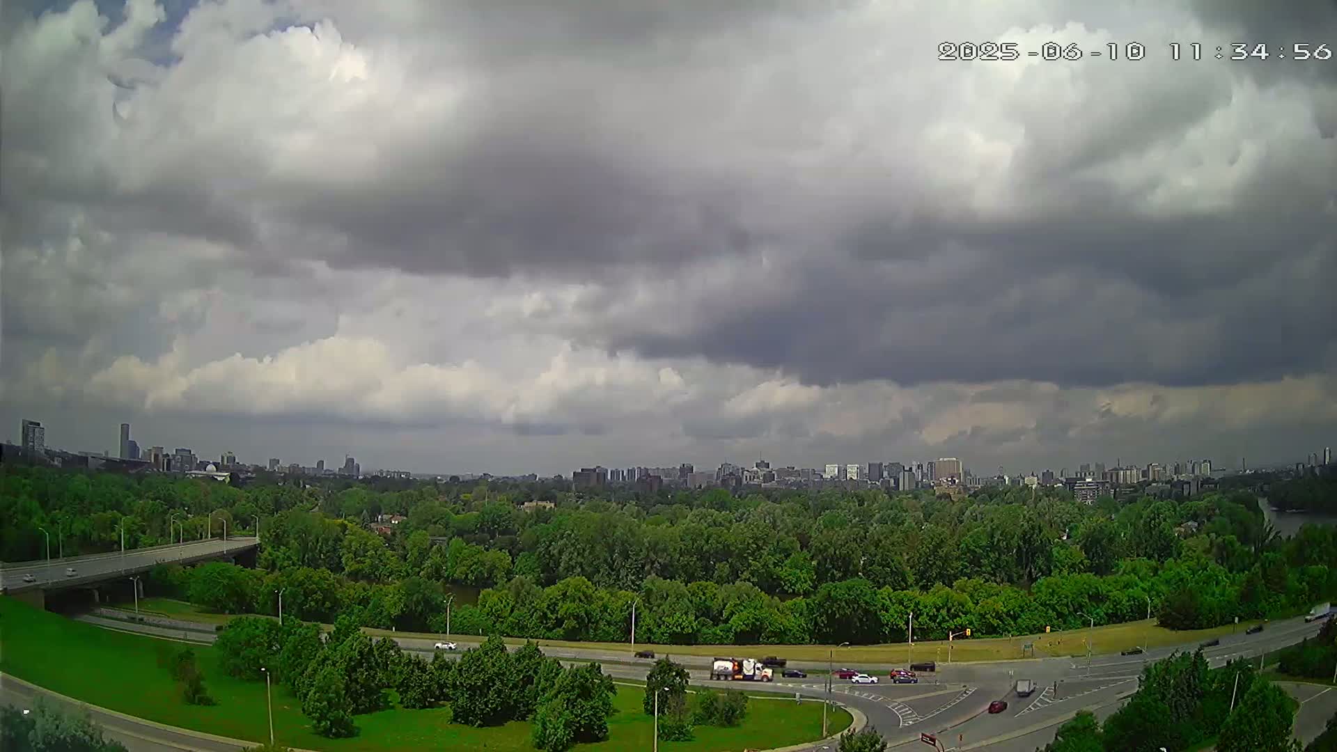 A highway curves through a lush green landscape under a mostly cloudy sky, with a city skyline visible in the distance.