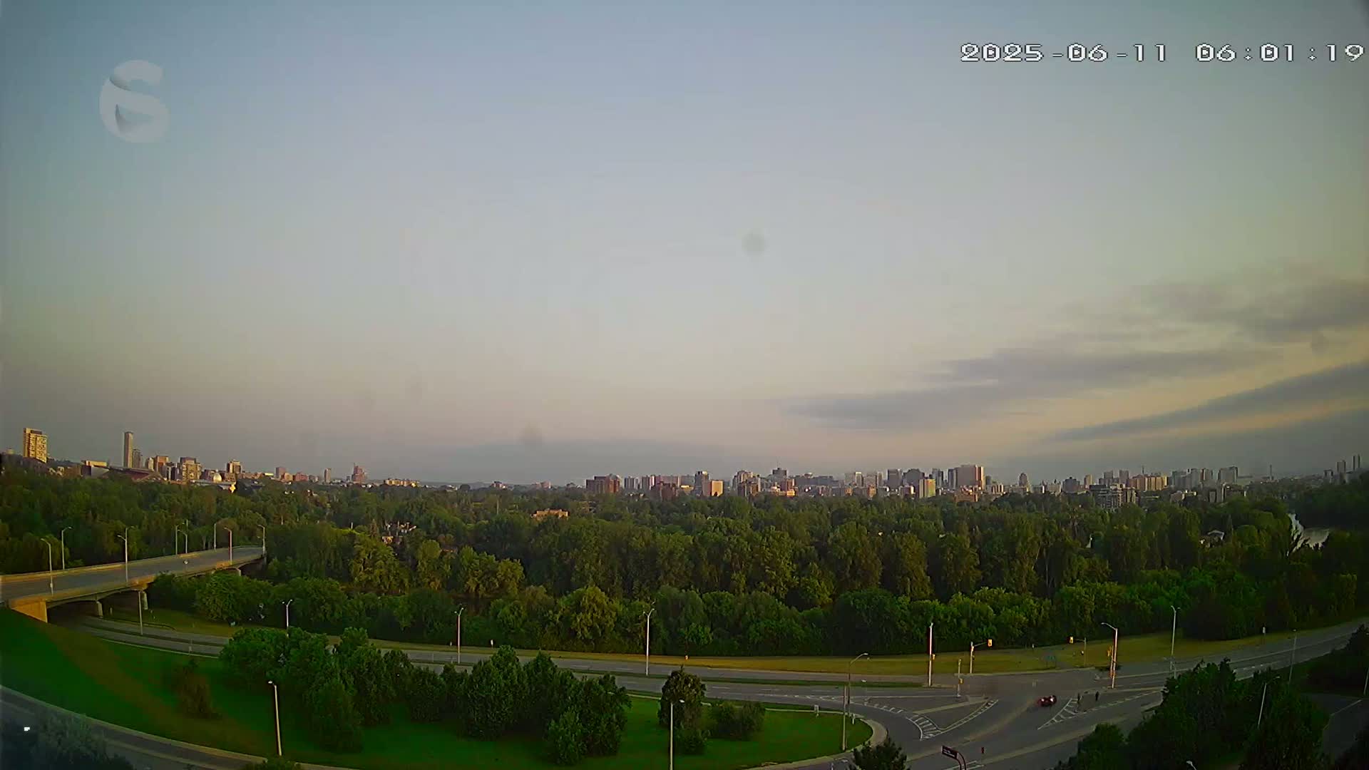 A city skyline is visible in the distance across a landscape of green trees and roads under a partly cloudy, light-colored sky.