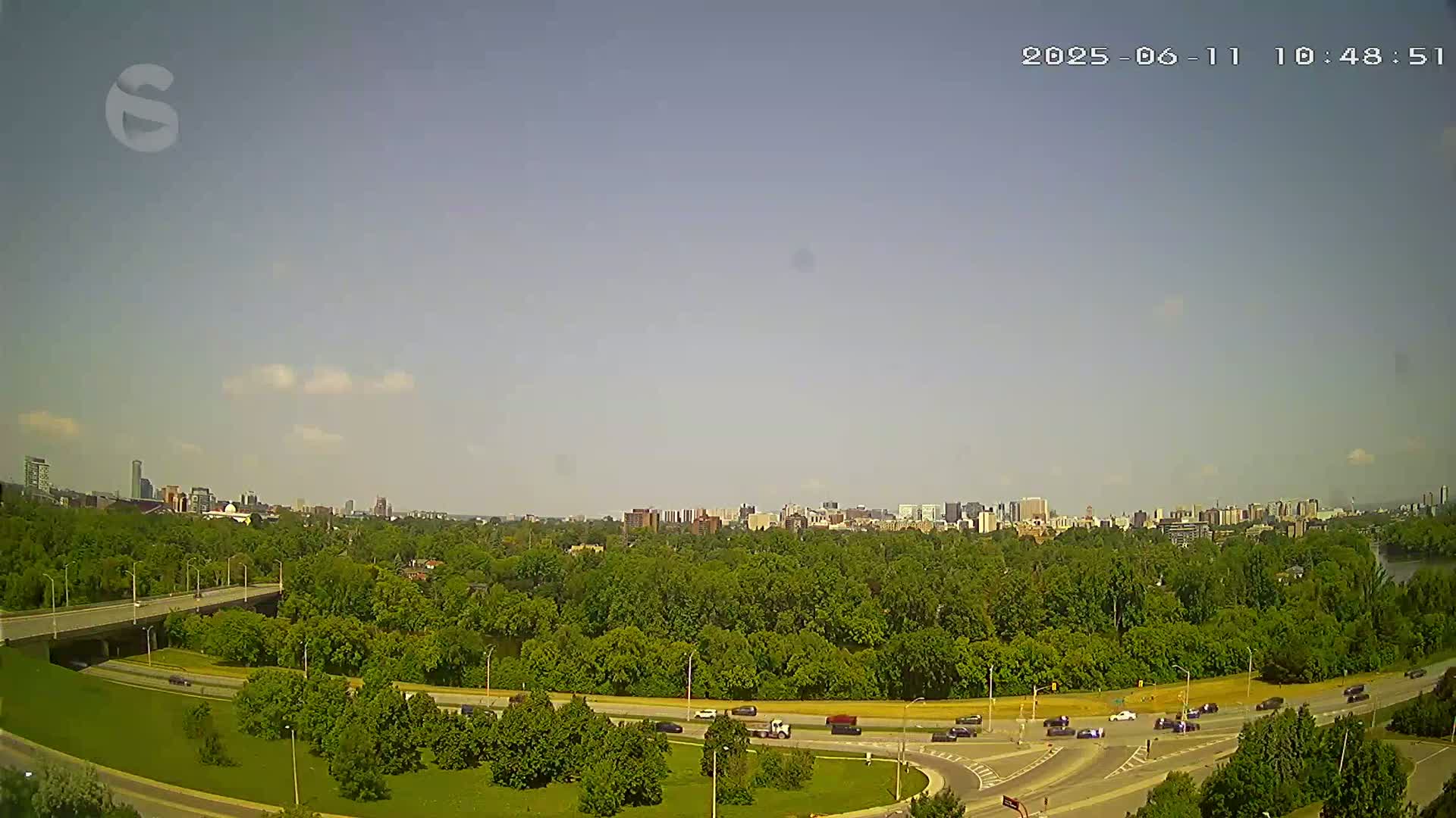 A highway curves through a lush green landscape under a mostly clear, sunny sky, with a city skyline visible in the distance.