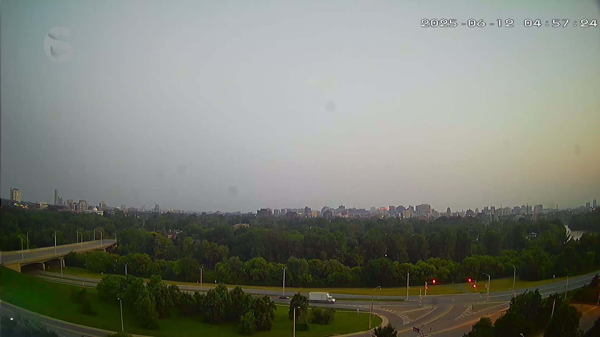 A hazy sky overlooks a highway interchange and a city skyline on the horizon, surrounded by lush green trees.
