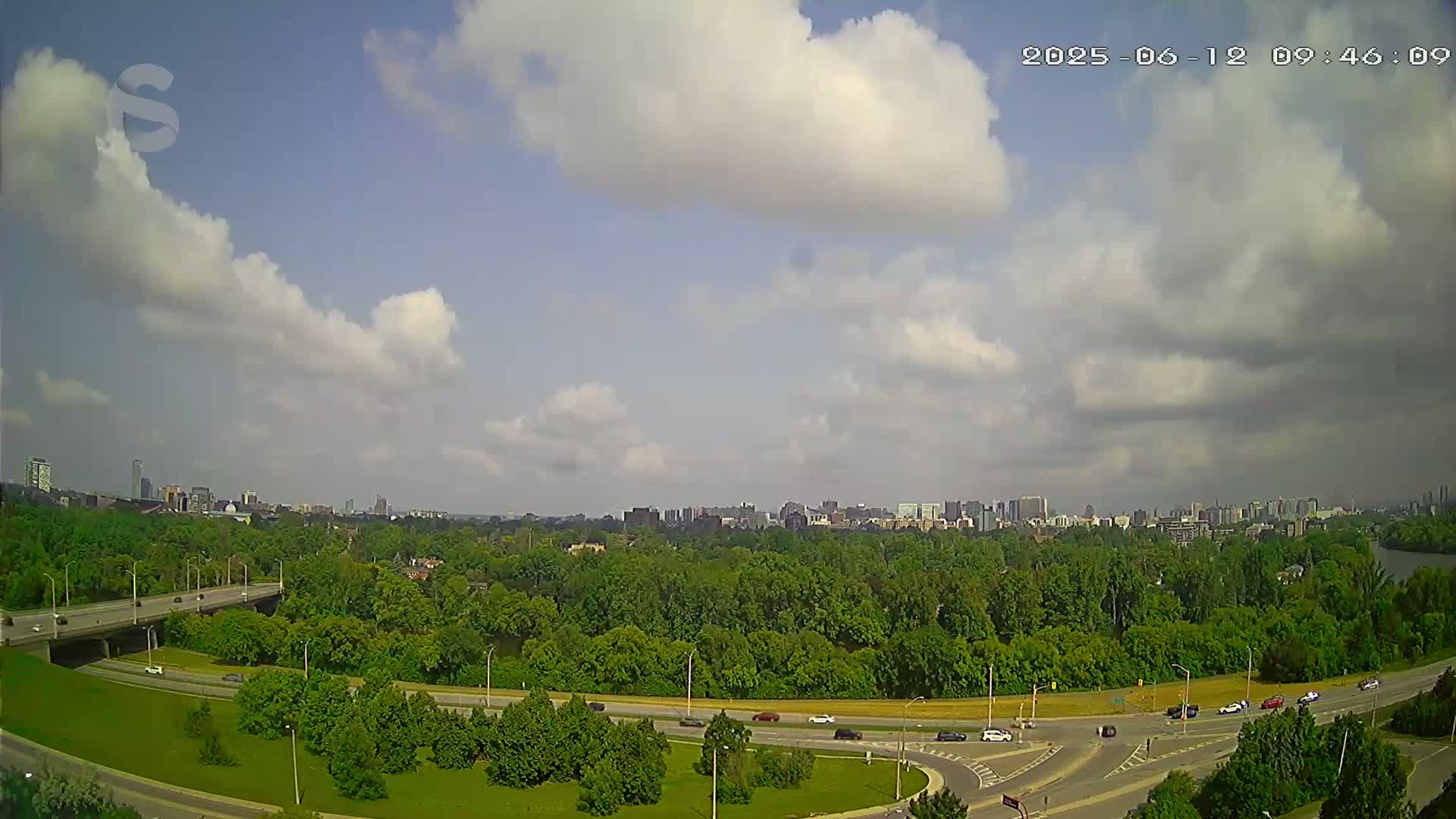 A partly cloudy sky overlooks a highway system curving through a lush green landscape, with a cityscape visible in the distance.