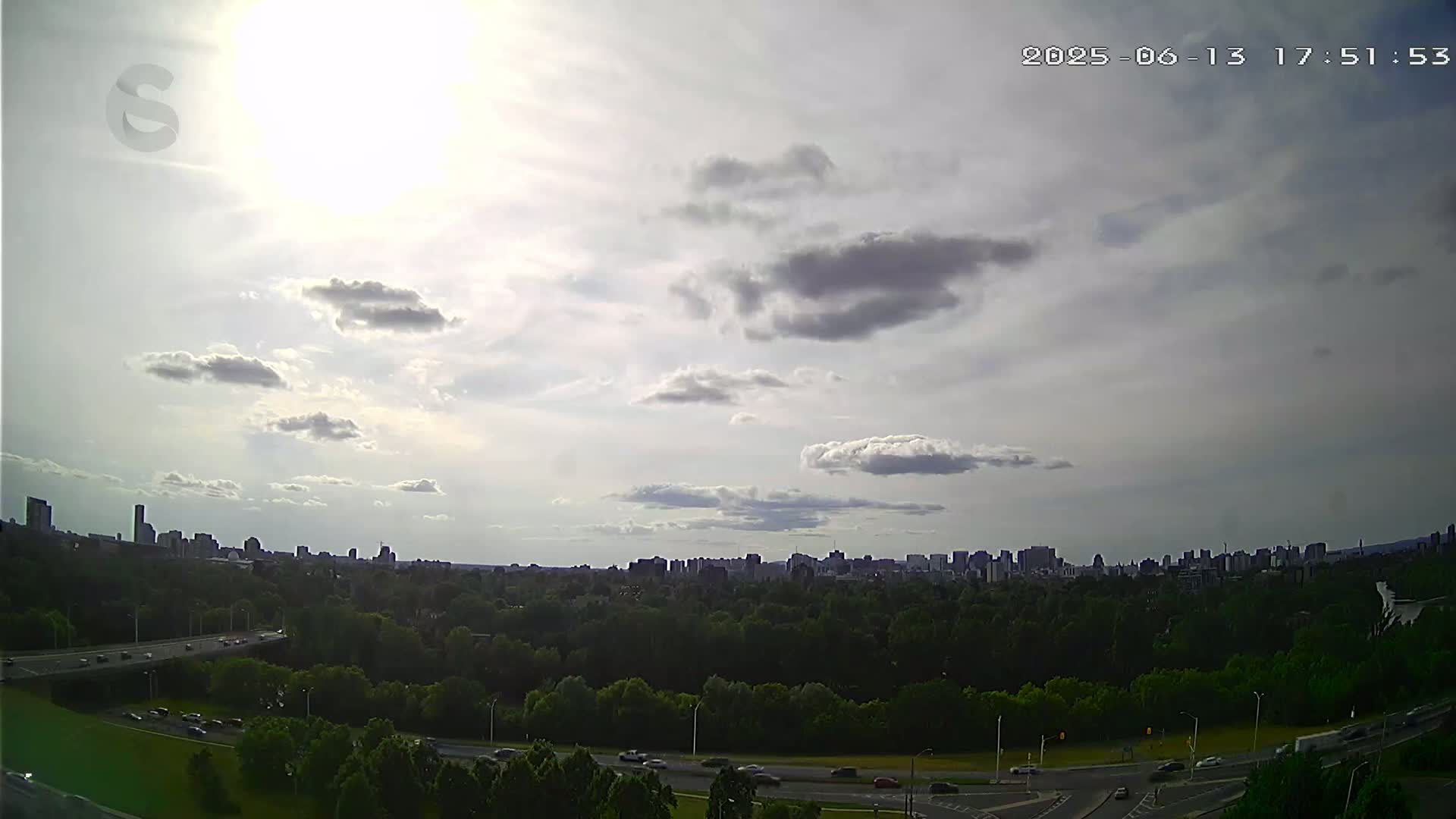 A partly sunny sky over a city skyline partially obscured by a lush green forest and a highway with sparse traffic.