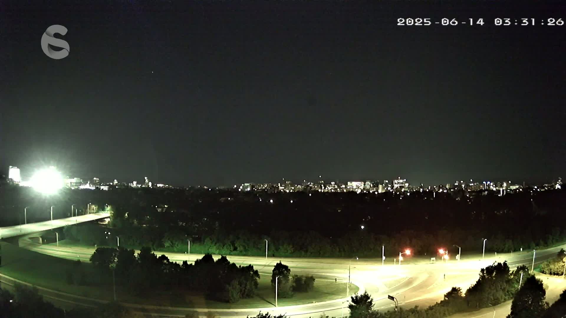 A nighttime view of a highway interchange with a city skyline in the distance under a clear sky.