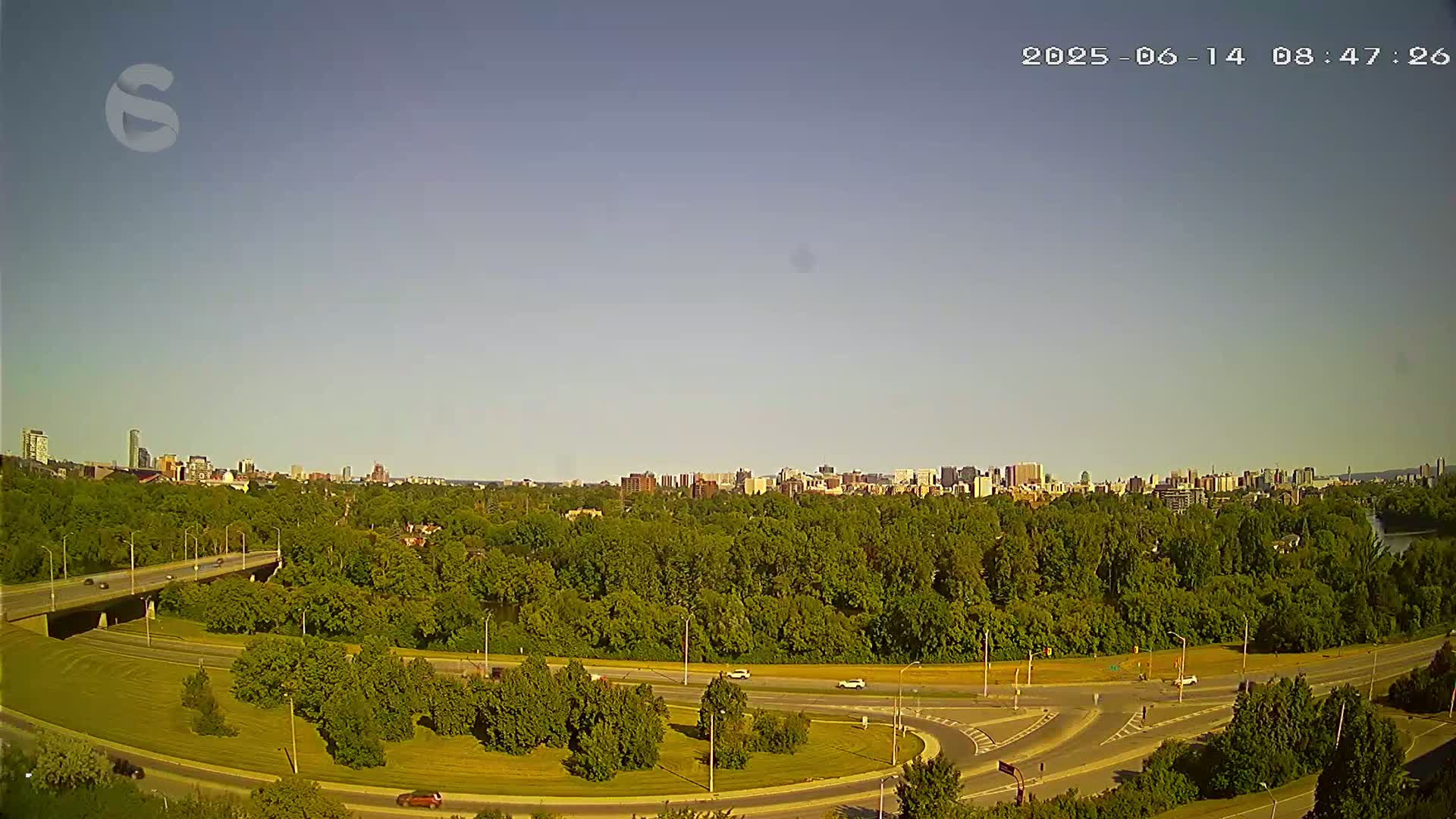 A highway curves through a green landscape, with a cityscape visible in the distance under a clear sky.