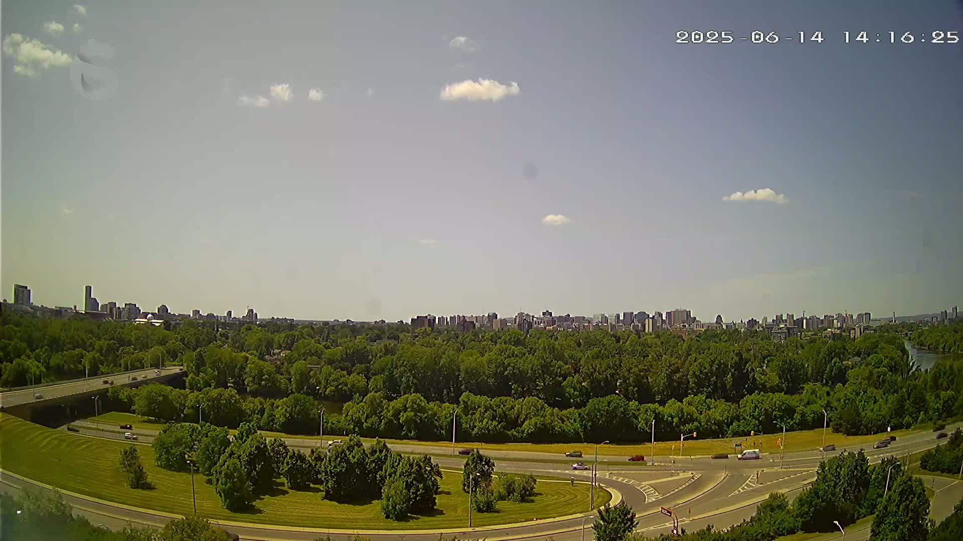 A city skyline is visible in the distance beyond a lush green park with a highway curving through it, under a mostly sunny sky with a few scattered clouds.