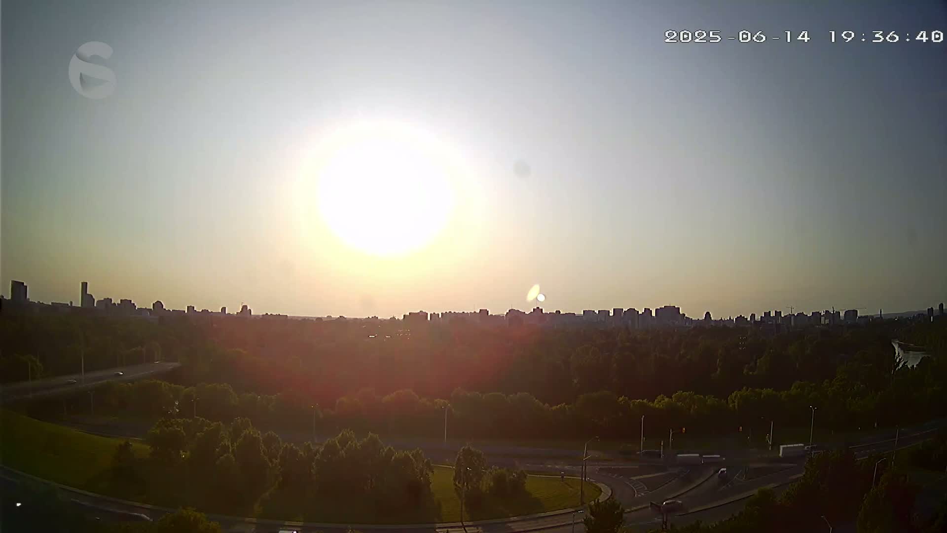 A city skyline is visible in the distance beyond a lush green park with a highway curving through it, under a mostly sunny sky with a few scattered clouds.