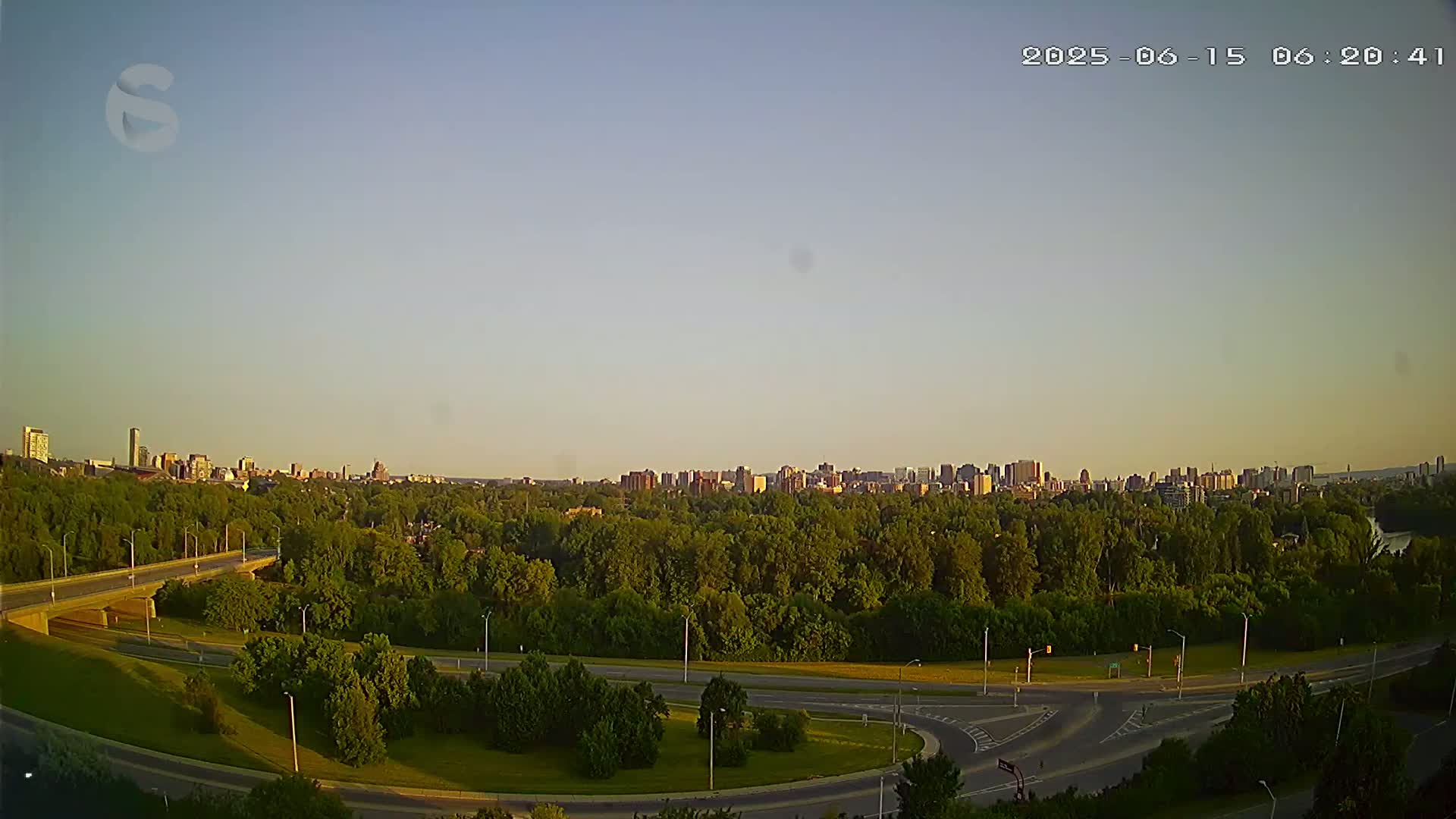 A city skyline is visible in the distance beyond a highway interchange and a large expanse of green trees under a clear, light-blue sky.