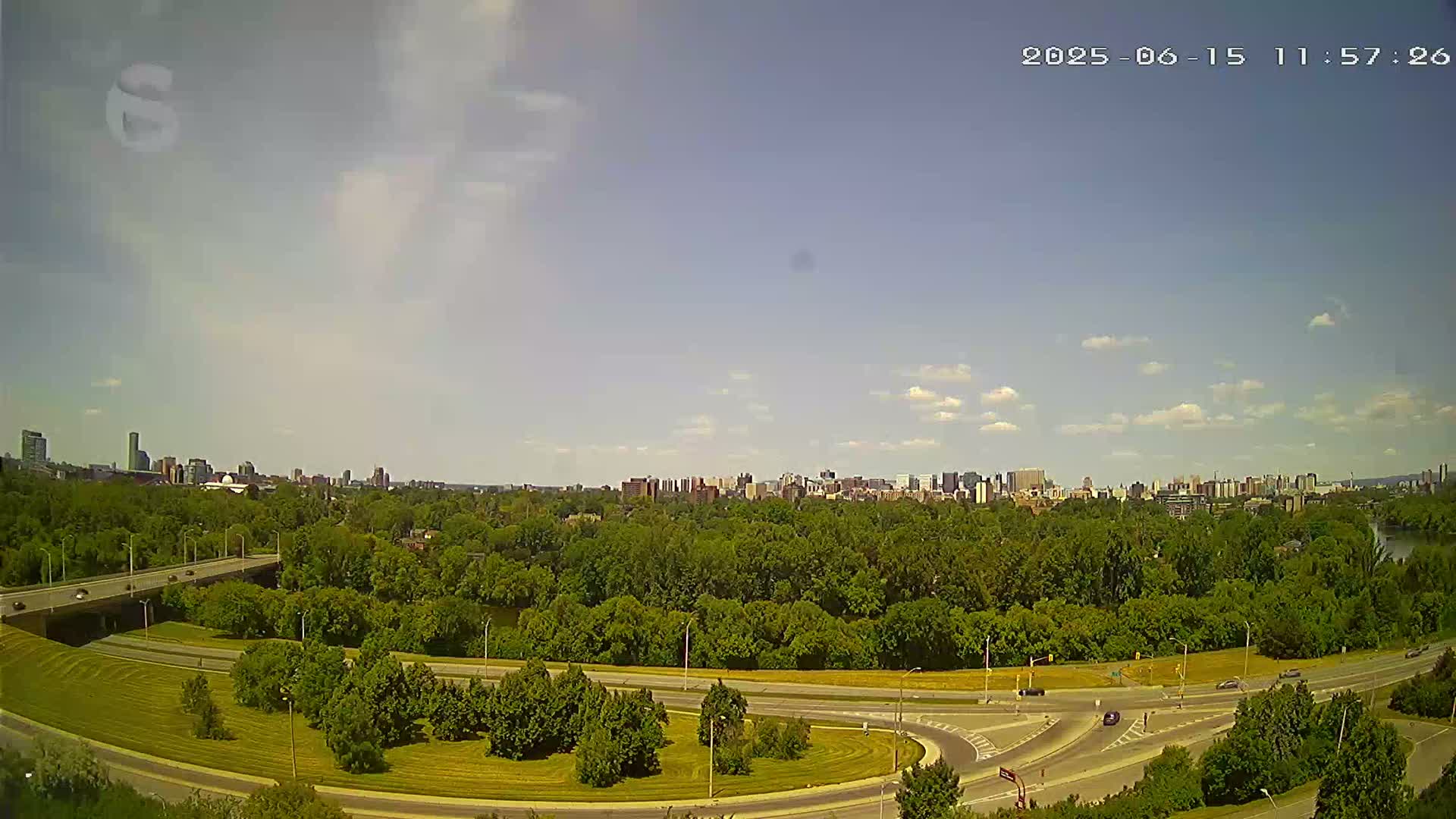 A city skyline is visible in the distance beyond a highway interchange surrounded by lush green trees under a mostly clear, sunny sky.