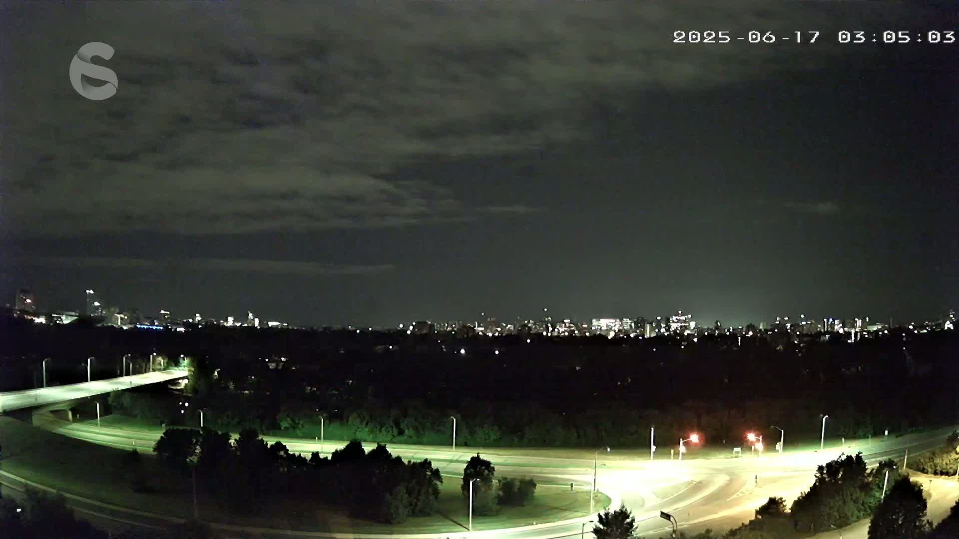 A nighttime view of a city skyline across a wooded area with a highway interchange below, under a mostly cloudy sky.