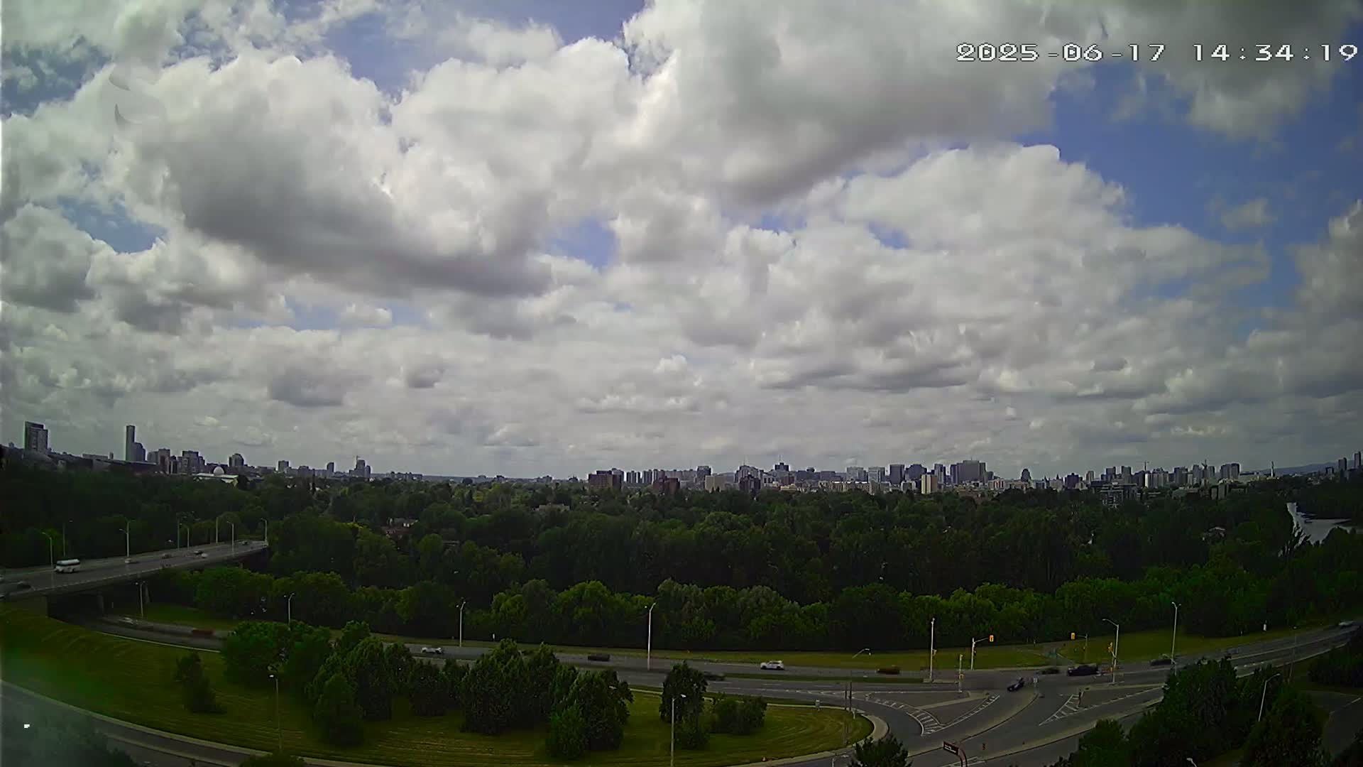 A partly cloudy sky overlooks a highway system curving through a green, tree-lined area before a distant cityscape.