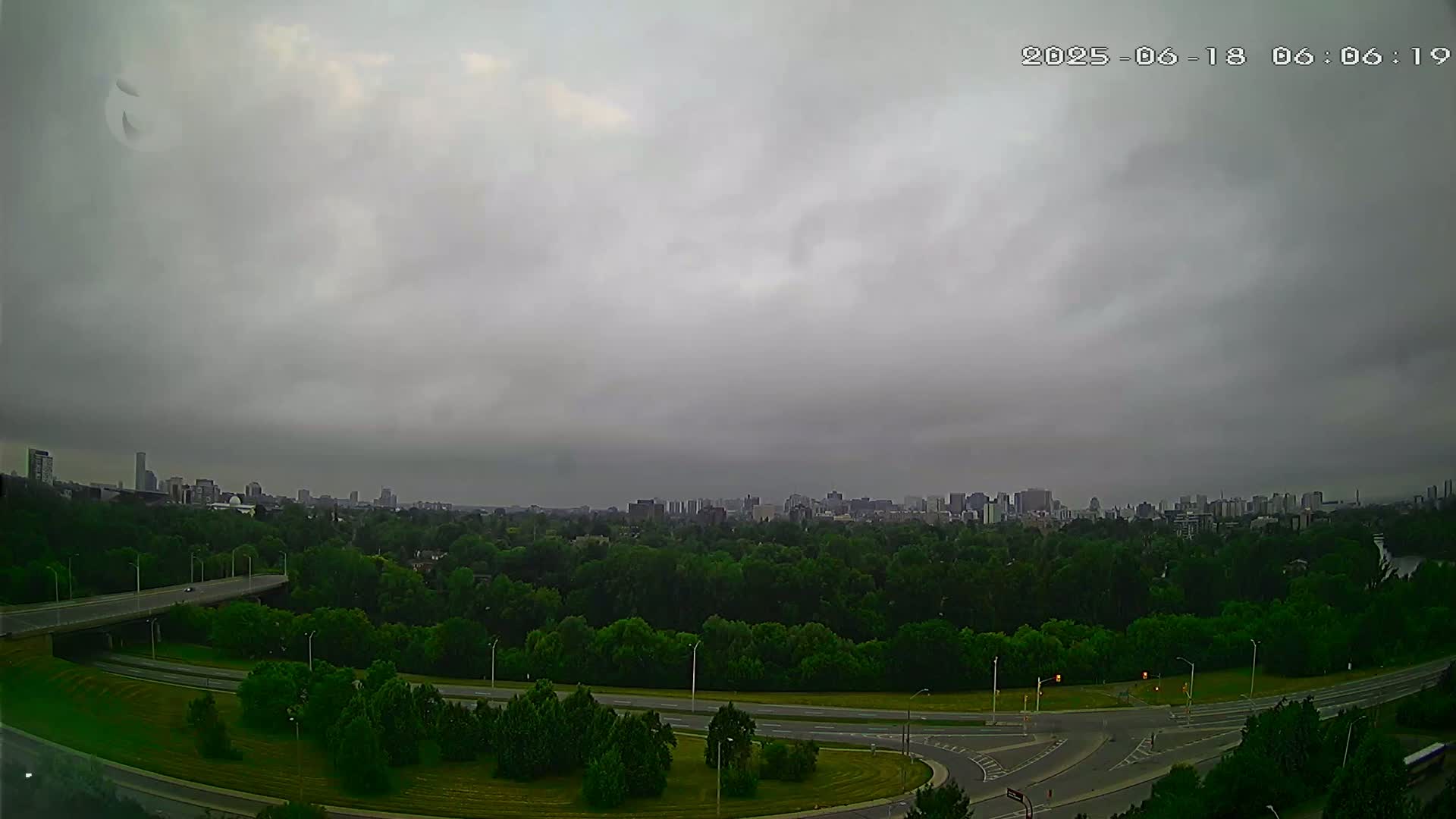 A city skyline is visible in the distance beyond a vast expanse of green trees and a curving highway under an overcast sky.