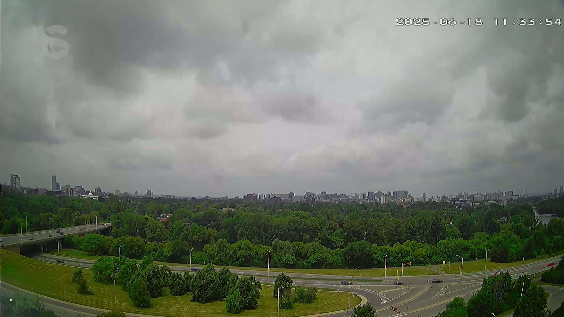 A highway curves through a lush green landscape under a cloudy sky, with a cityscape visible in the distance.