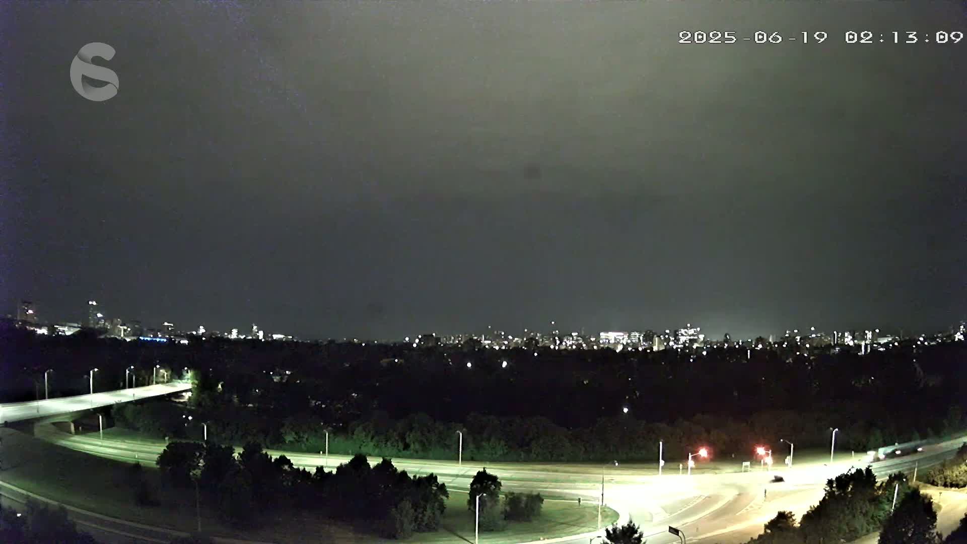 A nighttime view of a city skyline across a wooded area with a highway interchange below, under a cloudy sky.