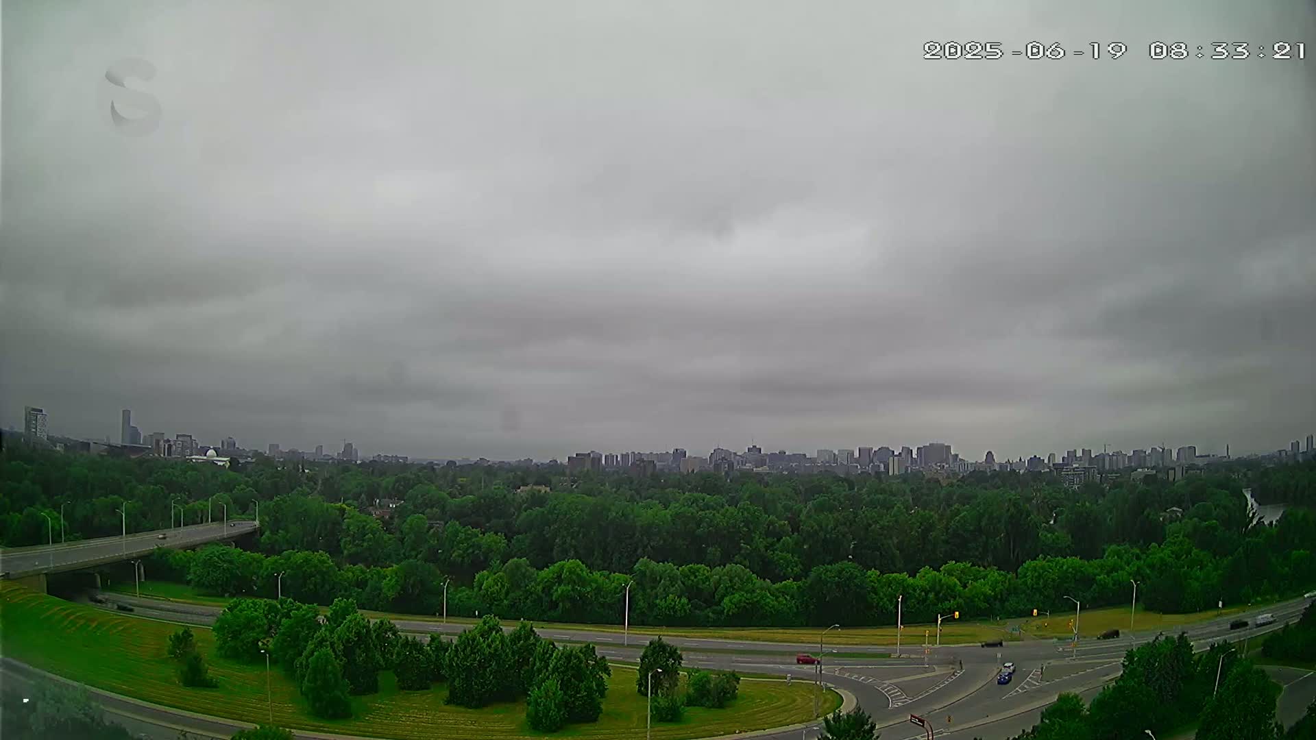 A city skyline is visible in the distance beyond a large expanse of green trees, a highway system, and overcast skies.