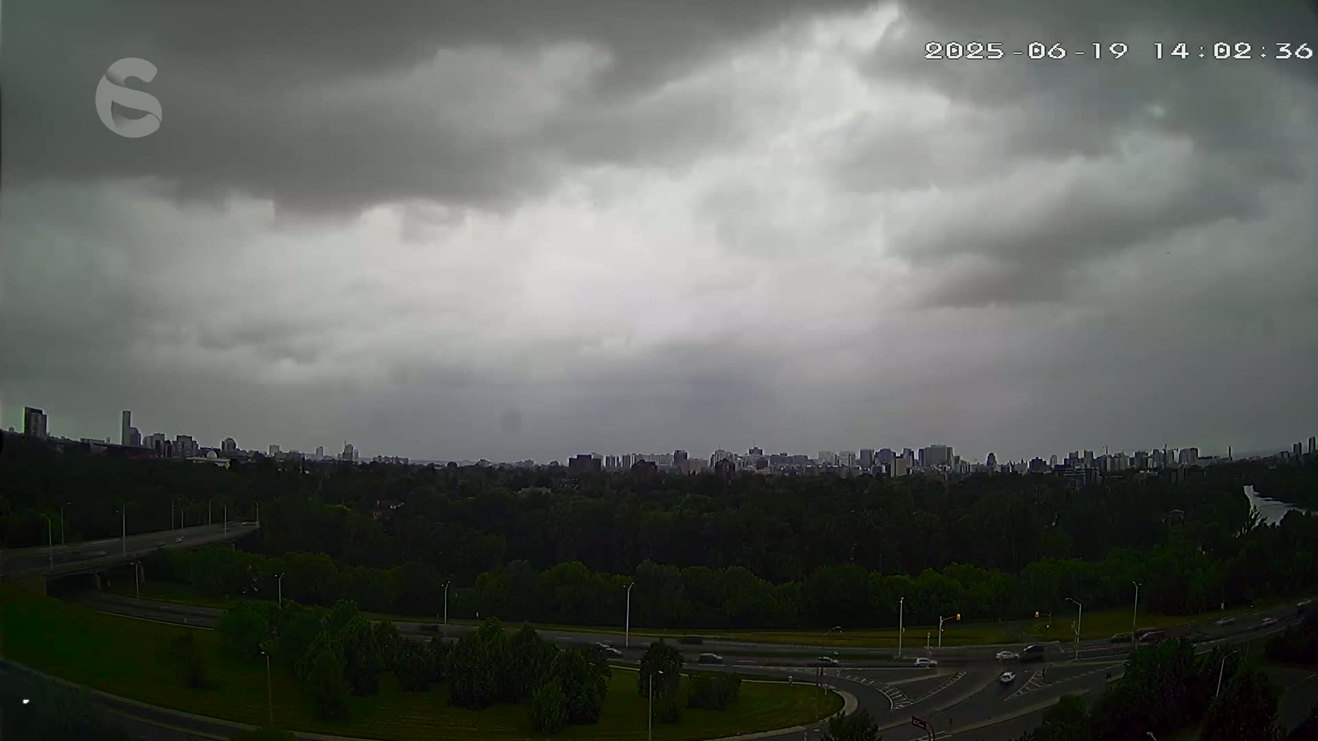 A city skyline is visible in the distance under a heavily overcast sky, with a highway and green space in the foreground.
