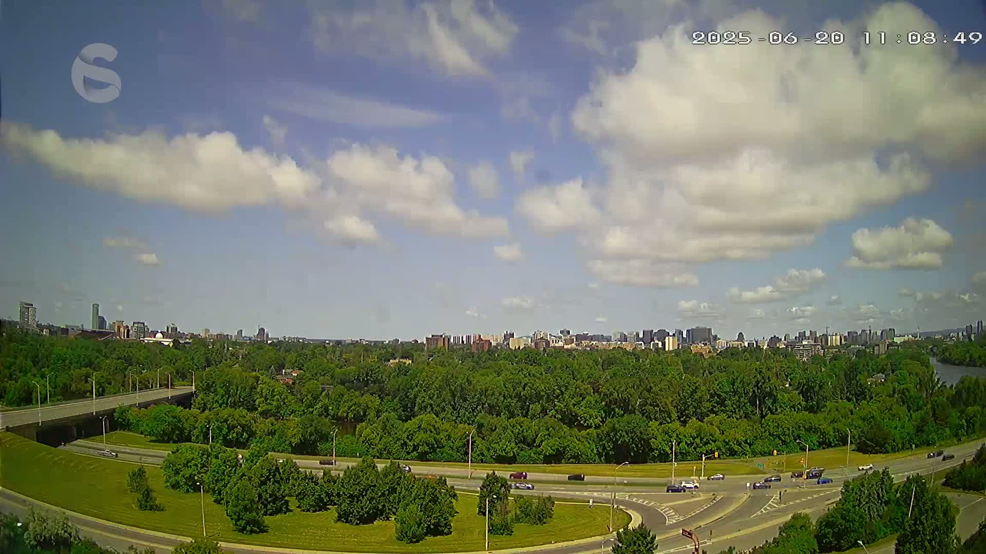 A partly cloudy sky overlooks a highway curving through a lush green landscape, with a city skyline visible in the distance.