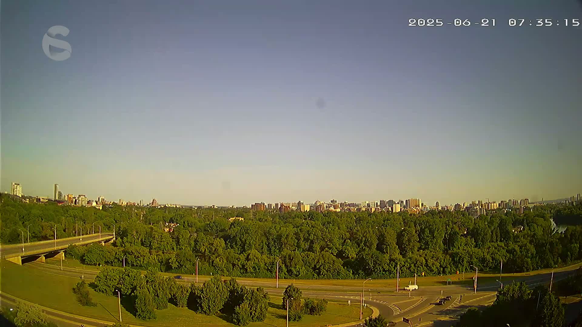 A clear, sunny day reveals a cityscape in the distance, partially obscured by a large expanse of green trees and a highway system with several interchanges.