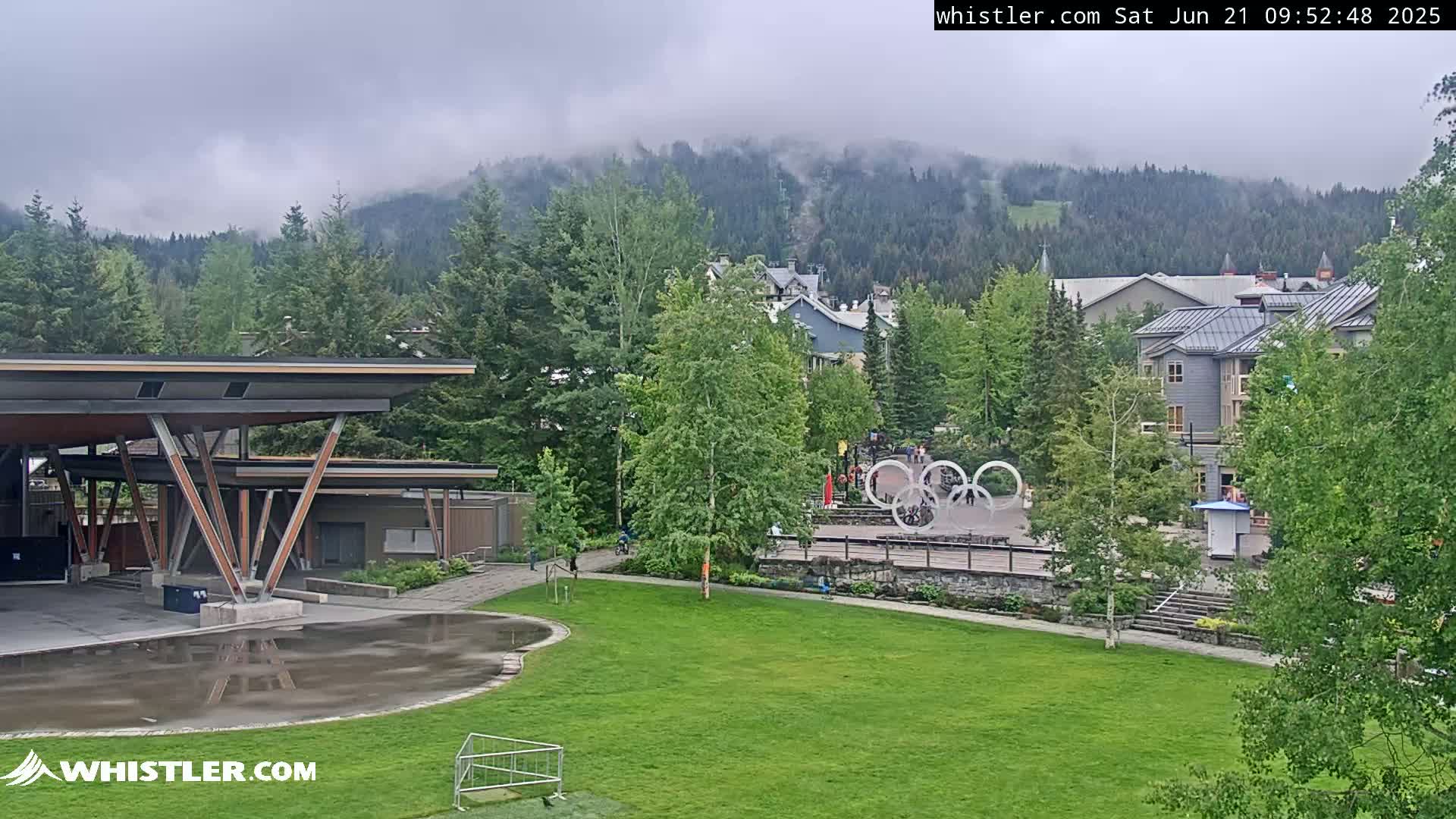 A cloudy view of a village nestled in a forested mountain valley, featuring a grassy park with an Olympic rings sculpture and a modern pavilion-like structure.