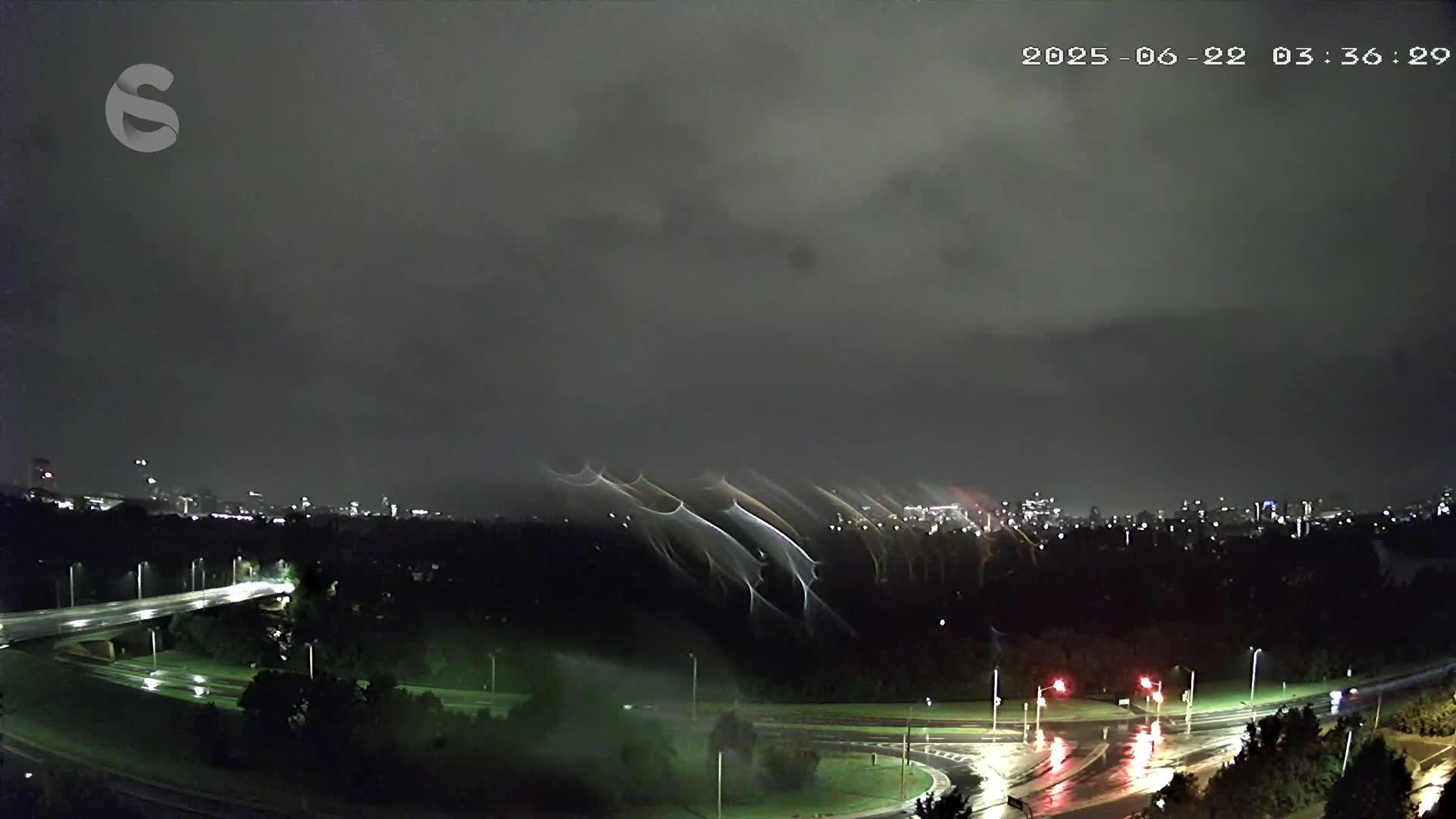 A nighttime view of a rain-slicked highway intersection and a cityscape in the distance under a dark, cloudy sky with unusual atmospheric phenomena visible above the trees.