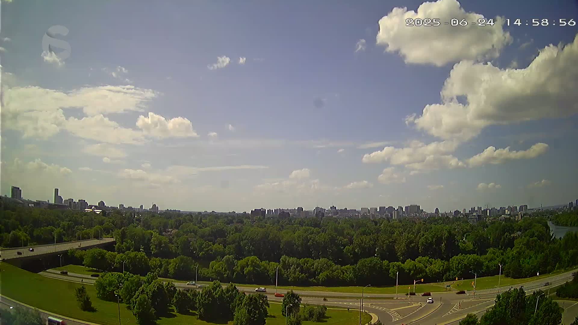 A partly cloudy sky overlooks a highway curving through a green landscape, beyond which a city skyline is visible in the distance.