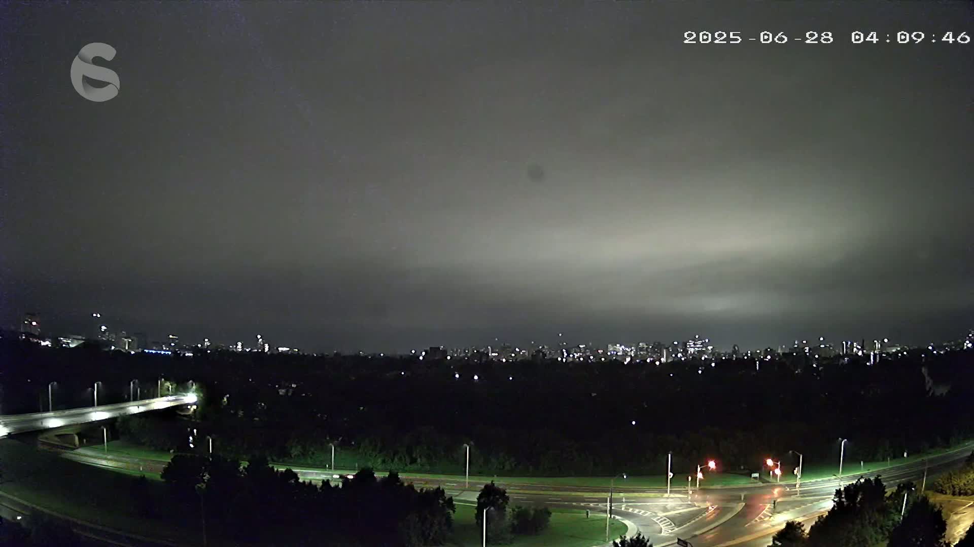 A nighttime, overcast view shows a city skyline in the distance beyond a dark, tree-lined roadway with illuminated traffic signals and a bridge.